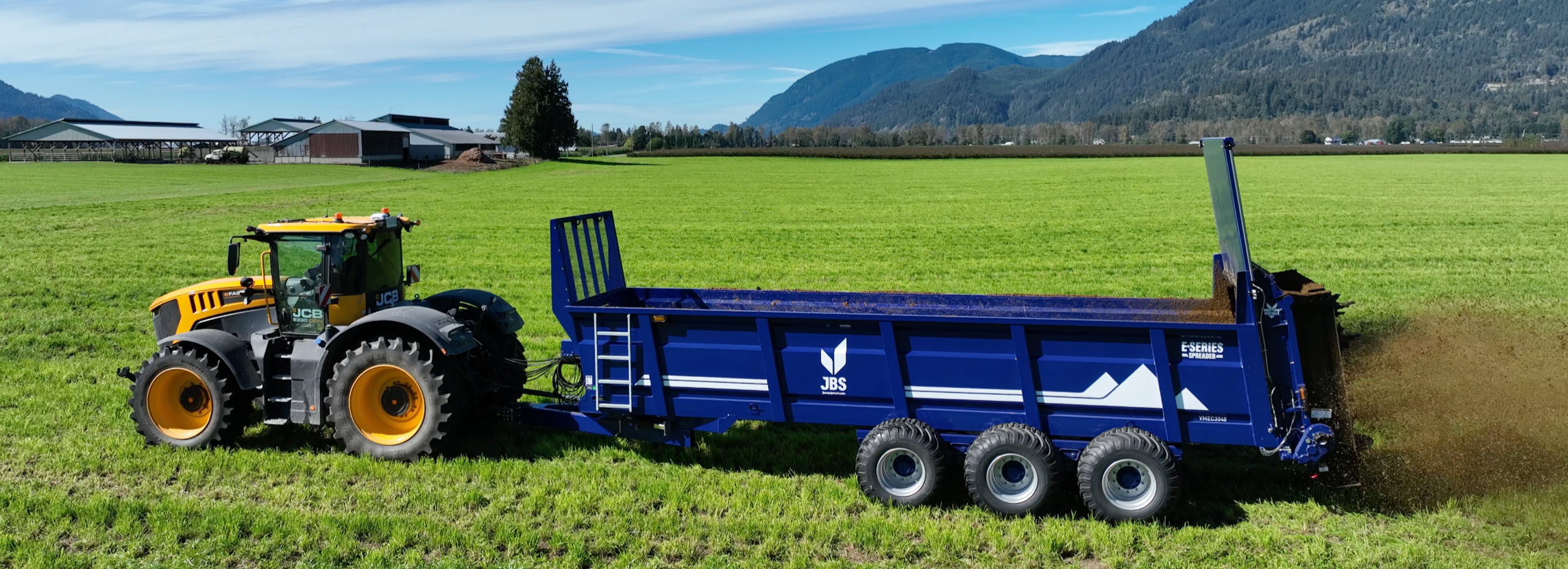 A yellow tractor with a blue trailer spreading fertilizer or soil on a green field with mountains in the background under a clear sky.