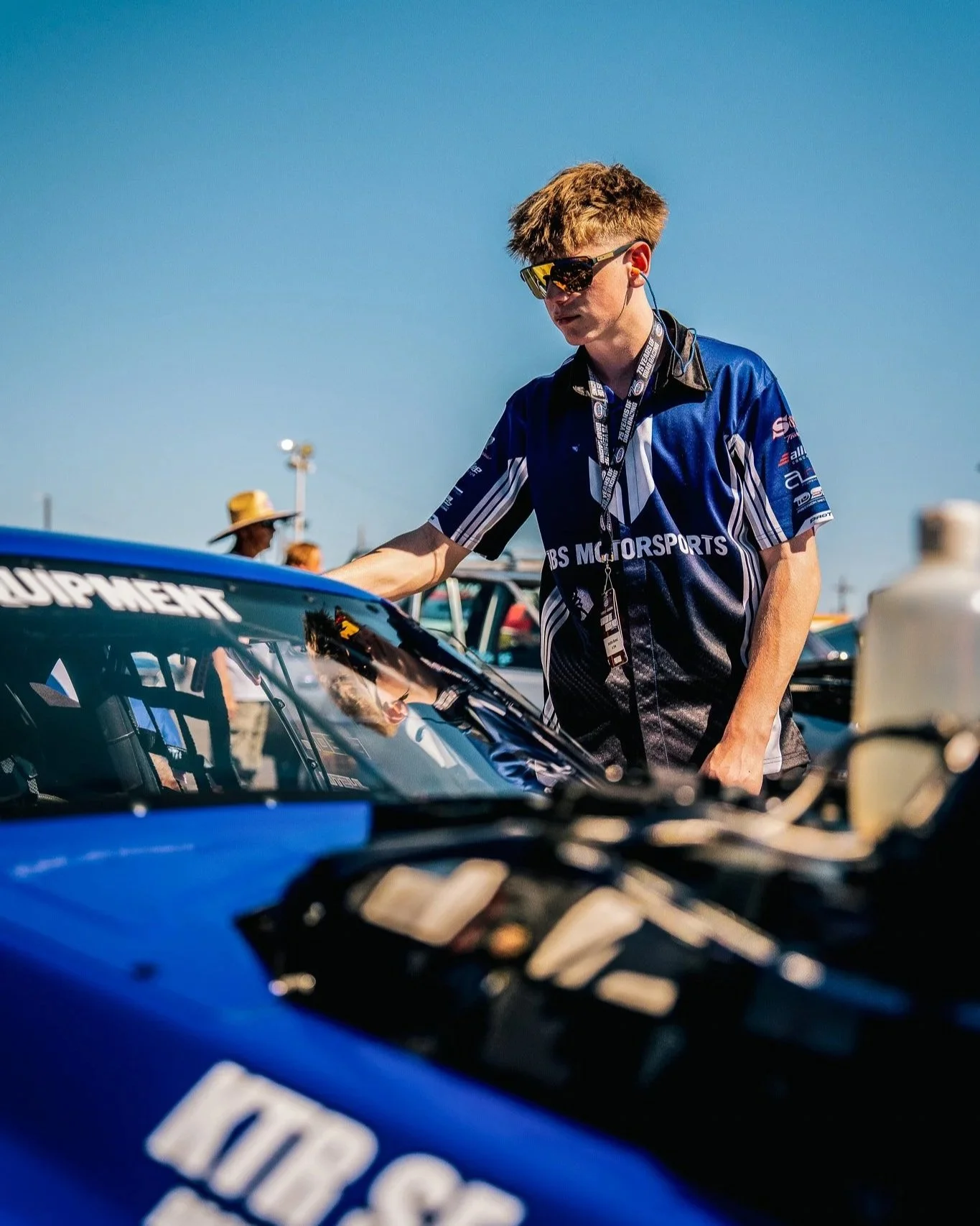 A young male race car driver with sunglasses and a team uniform, working on a blue race car in a sunny outdoor setting at a racing event.