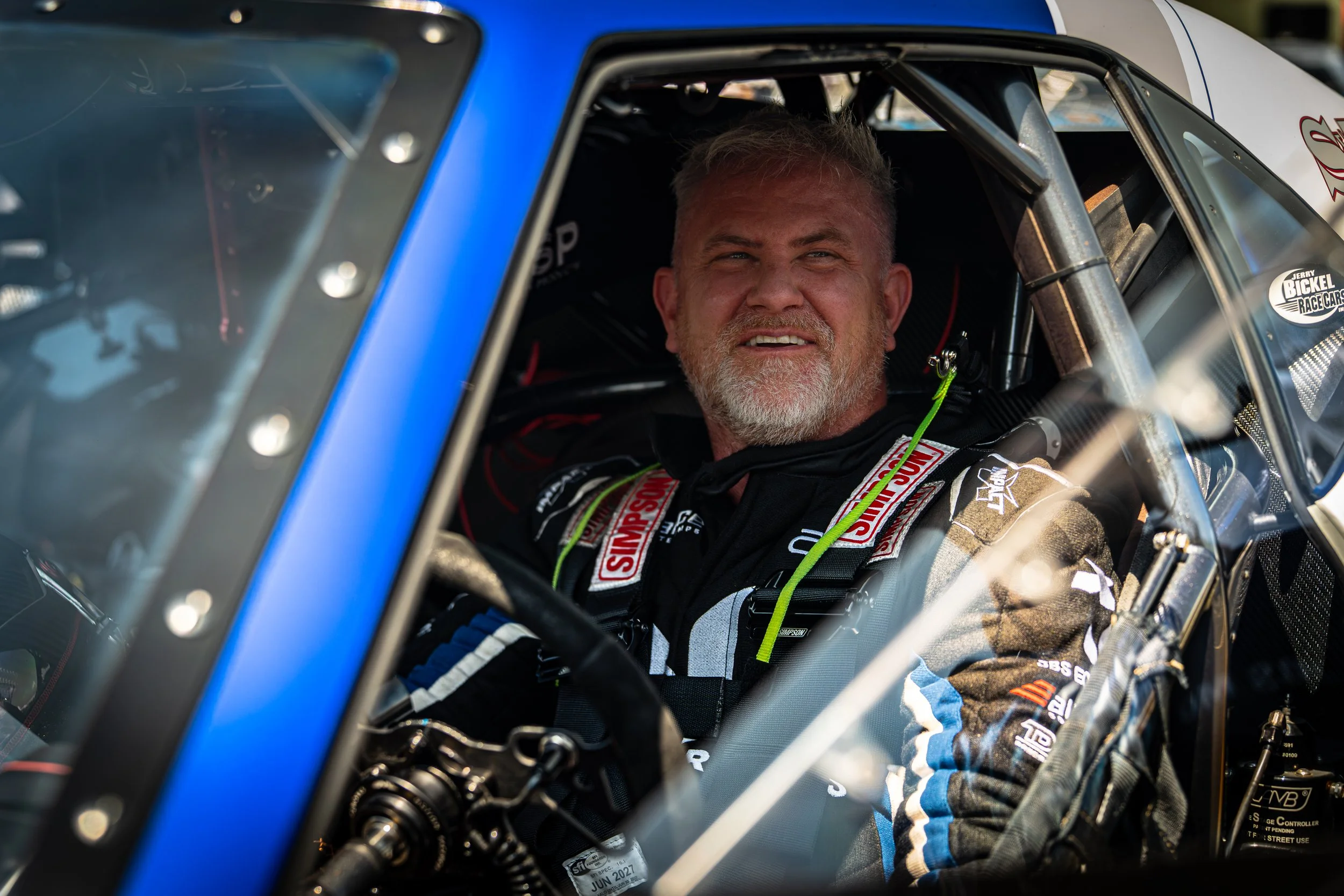 A race car driver sits inside his race car, smiling. The driver has a beard and gray hair, wearing a racing suit with various sponsor patches. The interior of the car includes safety harnesses and roll cage bars, with a window sticker indicating he is part of a racing event.