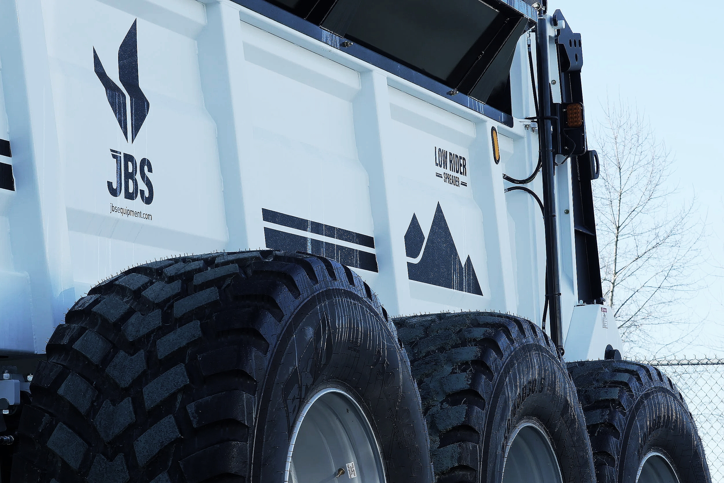 Close-up of large off-road tires on a white industrial vehicle with black equipment mounted on top, and logos reading JBS and Low Riser Spreader on the side, with clear blue sky and leafless trees in the background.