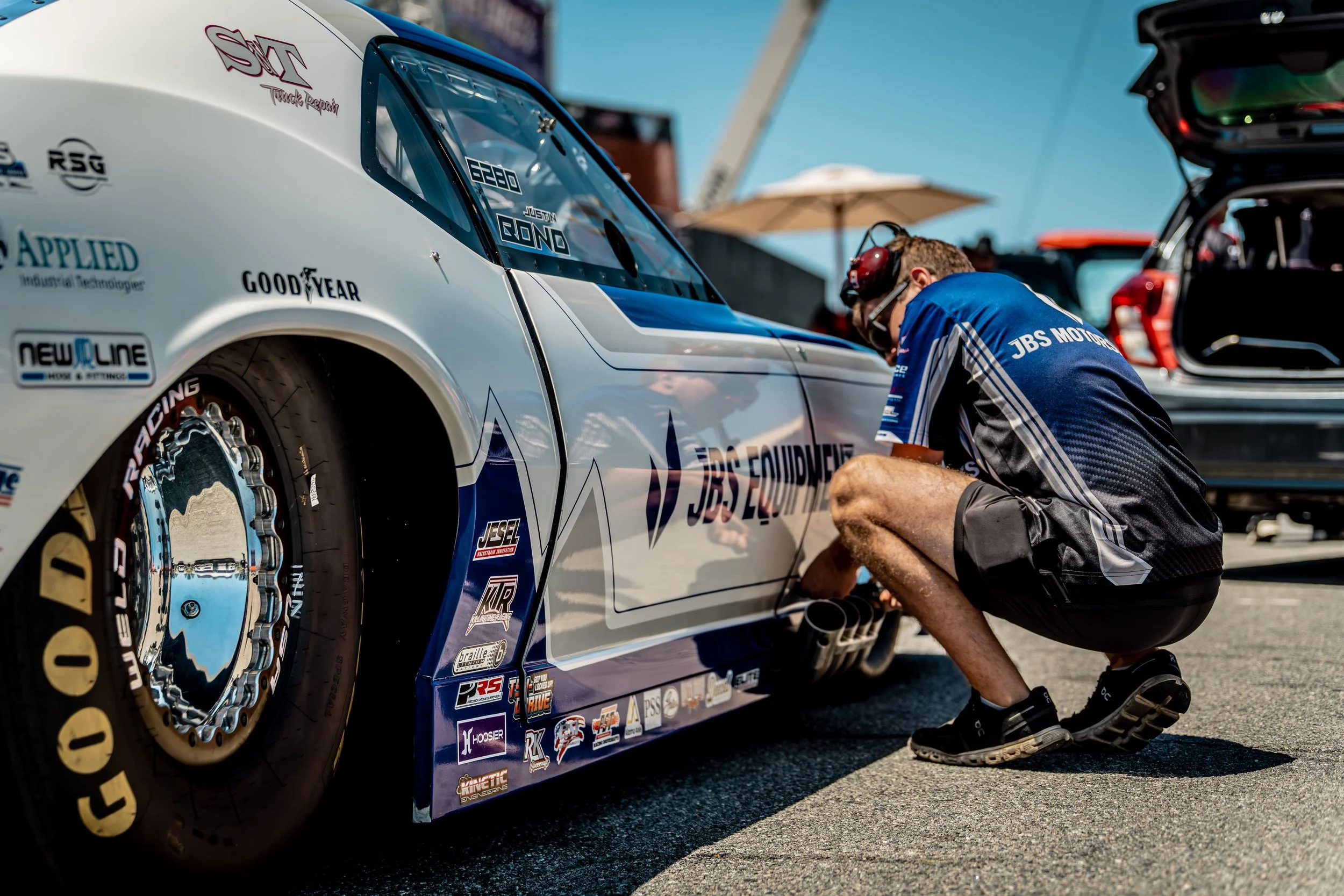A race car in a pit stop area with a mechanic working on the tire. The car is white with blue accents and various sponsor logos, including Goodyear tire branding. The mechanic is wearing a blue uniform and kneeling by the rear tire, which has a shiny, reflective rim.