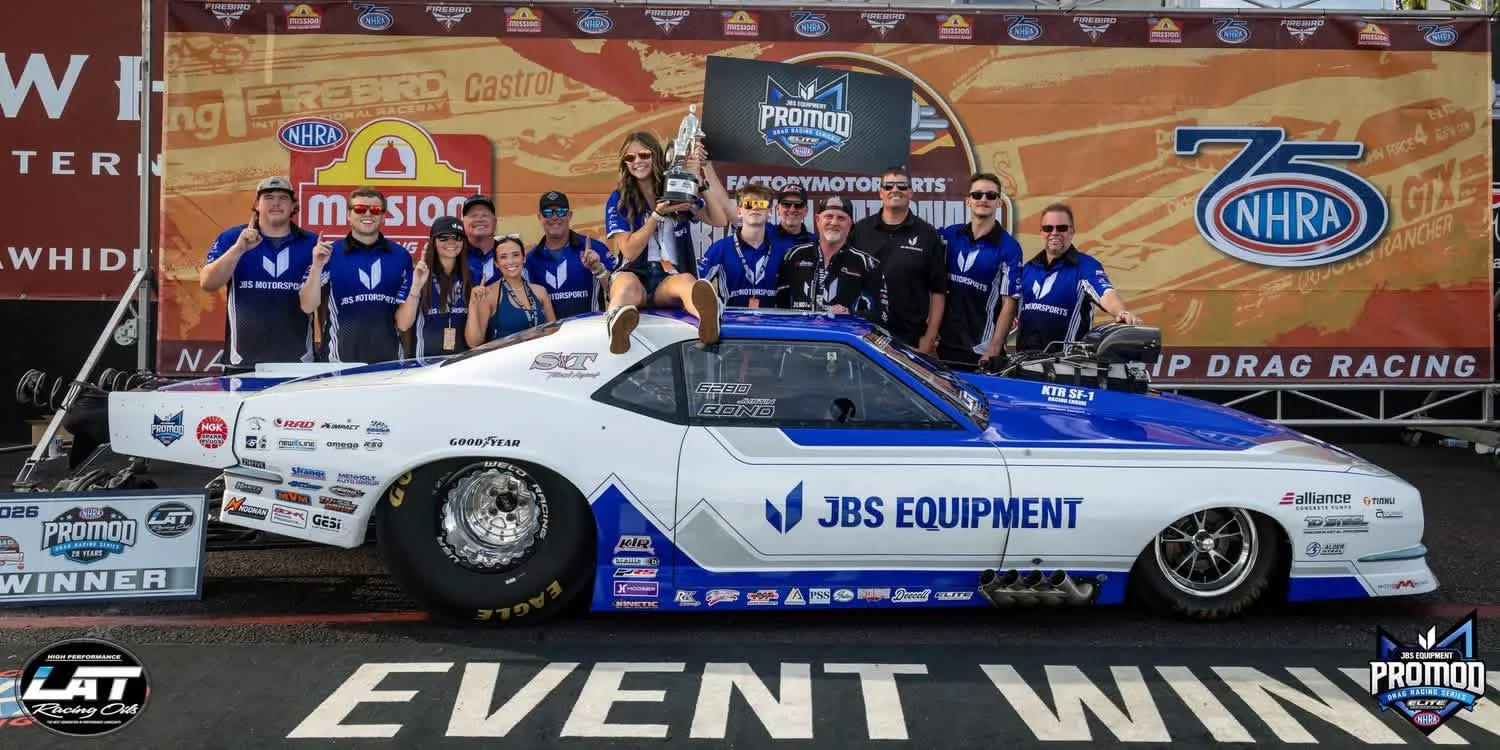 Group of people celebrating with a race car at the NHRA event, one person holding a trophy and sitting on the car, with banners and logos in the background.