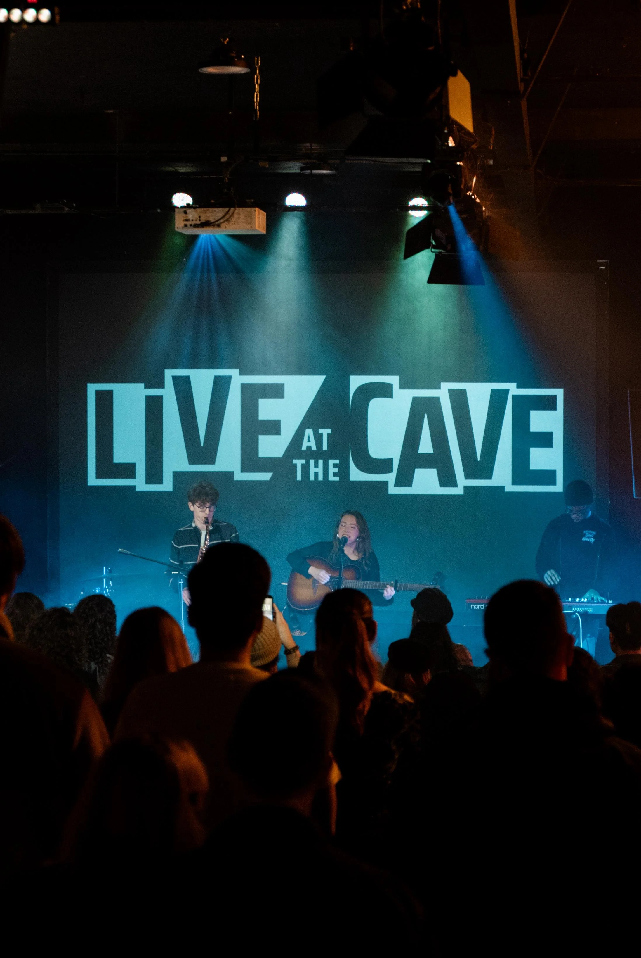 A live music performance on stage at the venue 'Live at the Cave.' A female musician is singing and playing guitar, accompanied by two other musicians, one playing saxophone and the other on keyboard. The audience is watching in the foreground.