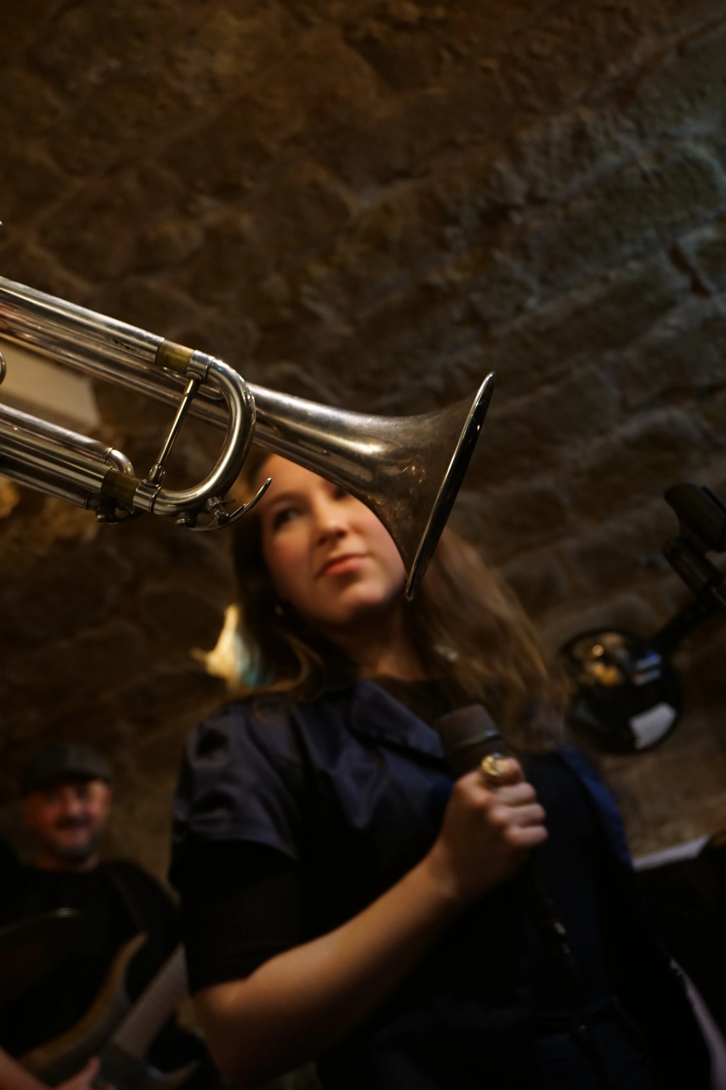 A woman holding a microphone and looking at a trumpet, with a man playing guitar in the background, in a room with a brick wall ceiling.