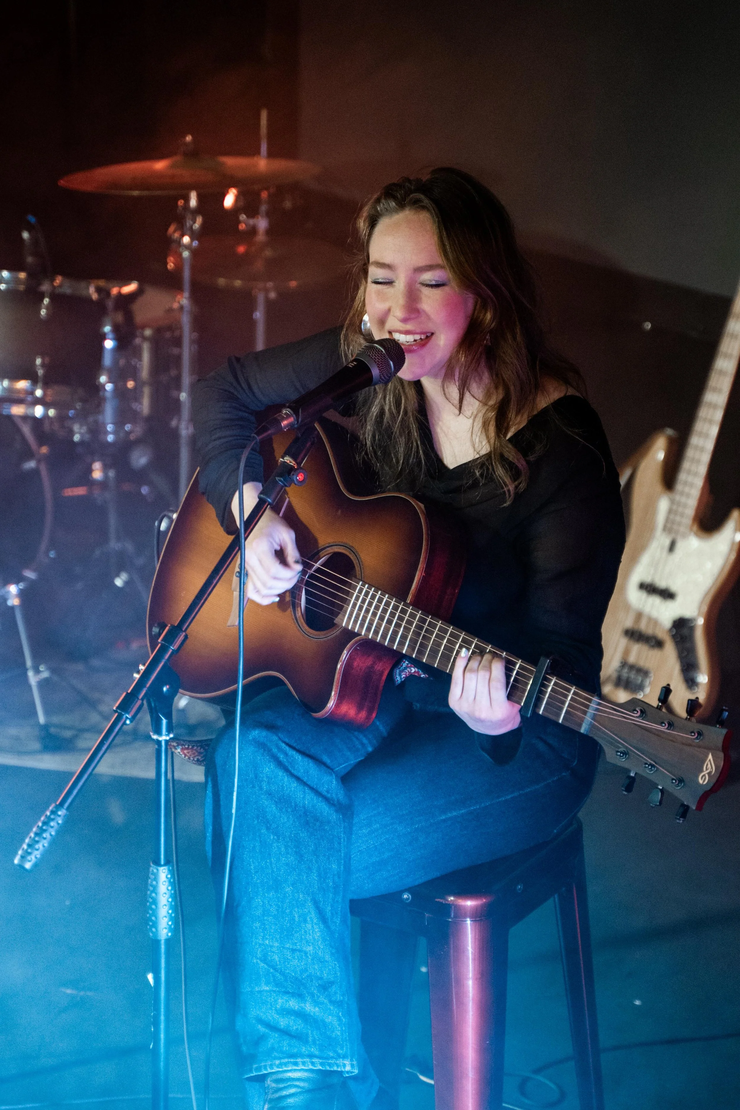 A woman sitting on a stool playing an acoustic guitar and singing into a microphone, with drums and an electric bass in the background.