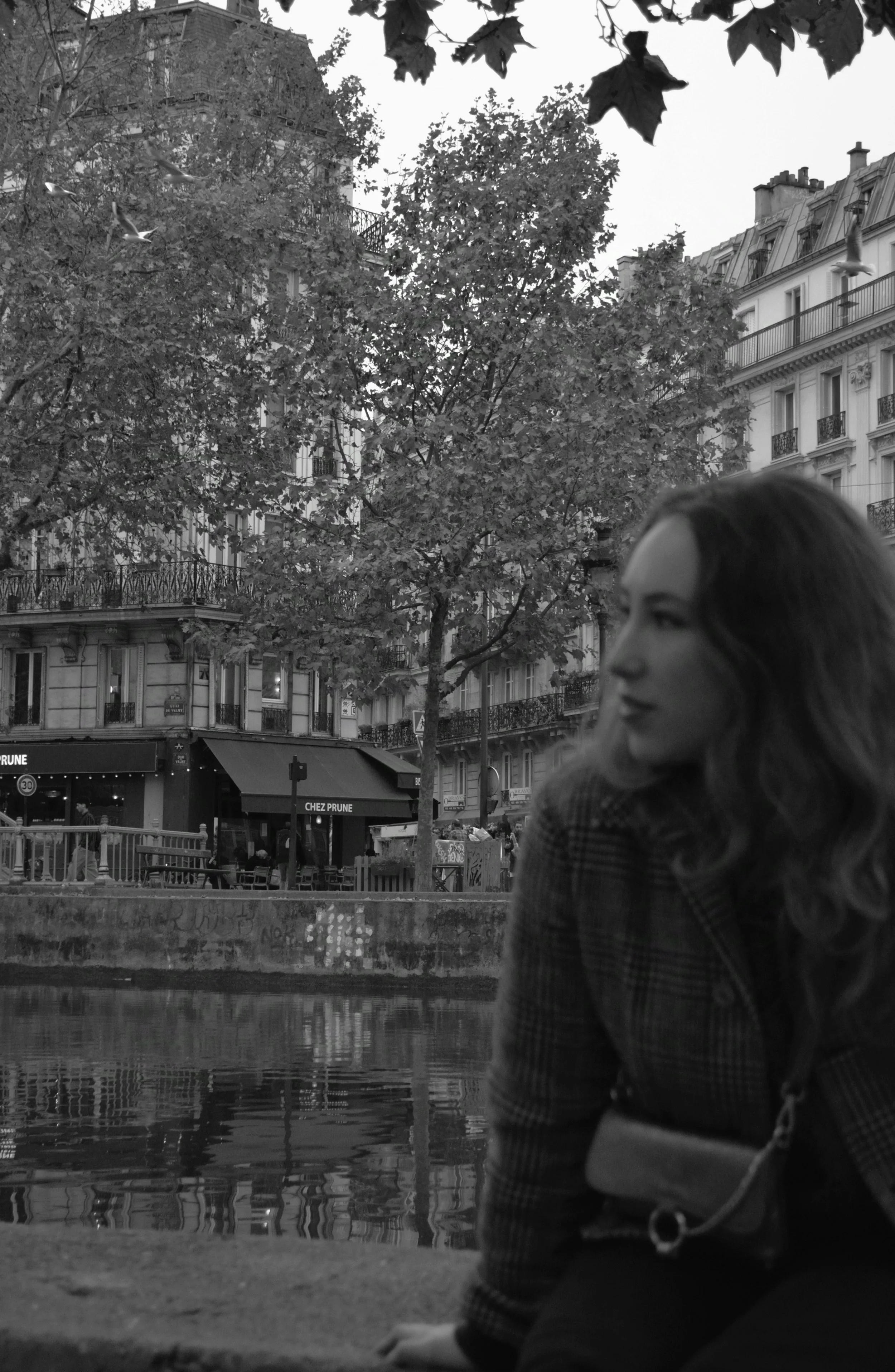 A woman with wavy hair sitting by a canal in a city, with trees and classic European-style buildings in the background, in black and white.