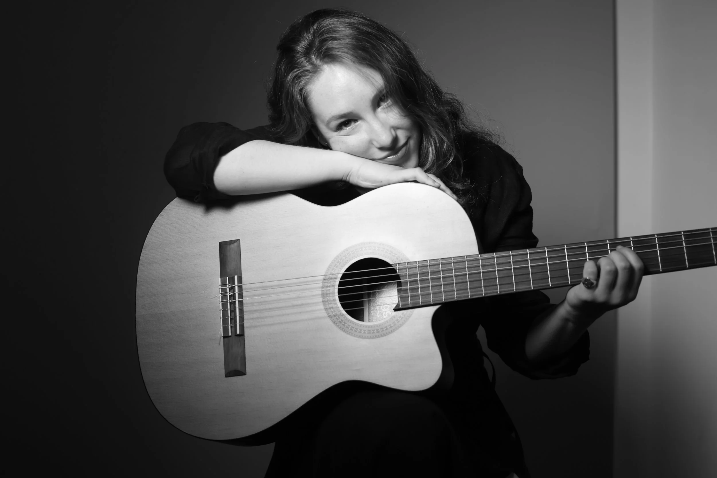 A young woman with curly hair smiling and resting her head on an acoustic guitar, black and white photo.