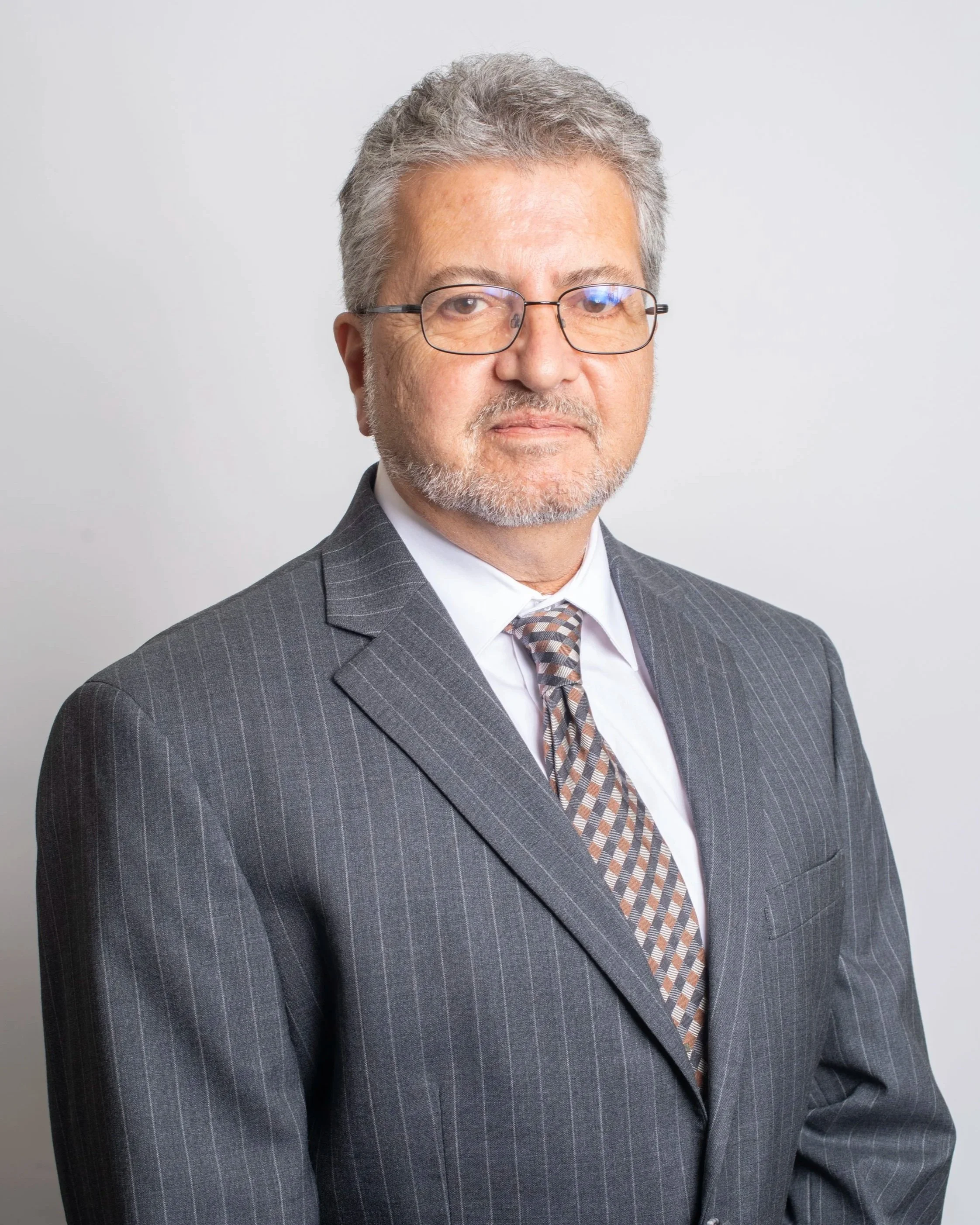 A middle-aged man with gray hair, wearing glasses, a gray pinstripe suit, a white shirt, and a checkered tie, stands against a plain white background.