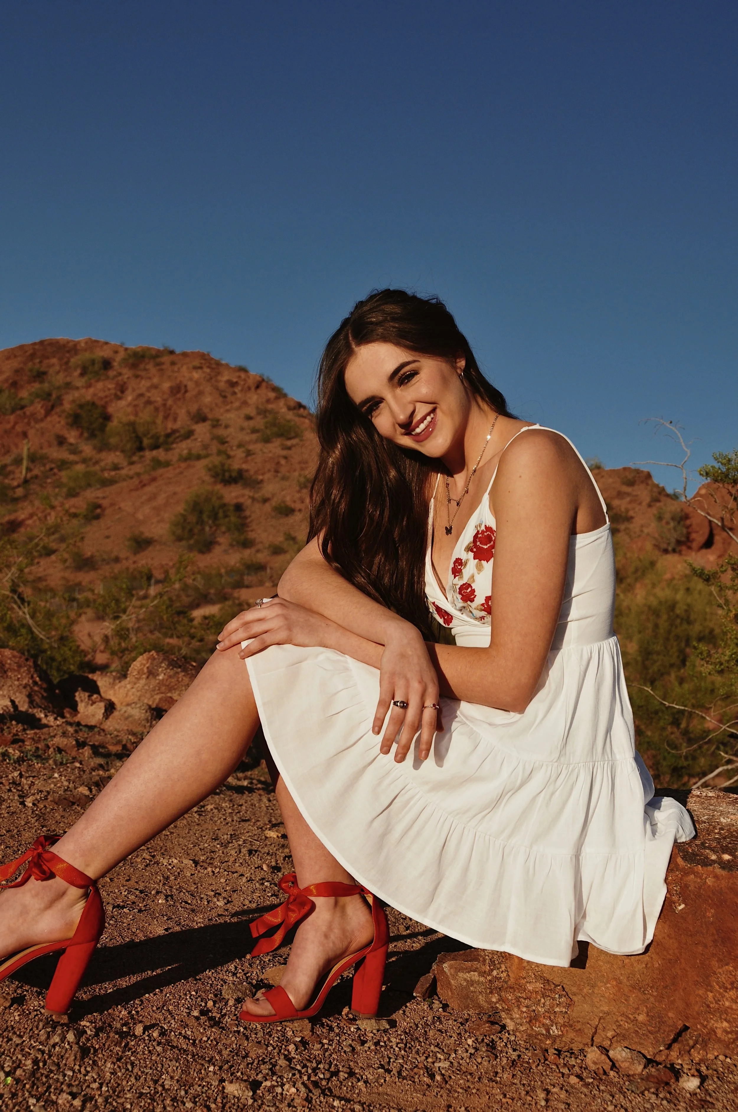 A young woman with long dark hair smiling while sitting outdoors on rocky terrain with desert hills and a clear blue sky in the background, wearing a white dress with floral embroidery, red high heels with ribbons, and gold jewelry.