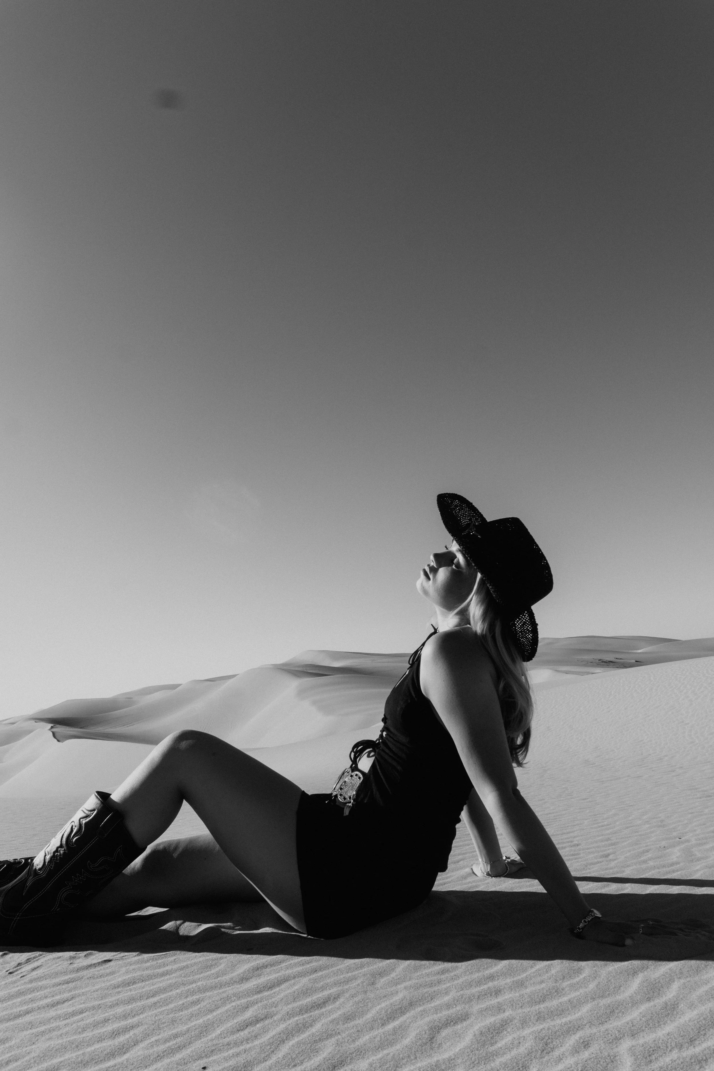A woman wearing a wide-brimmed hat, sunglasses, and a dress, sitting on sand dunes in a desert landscape.