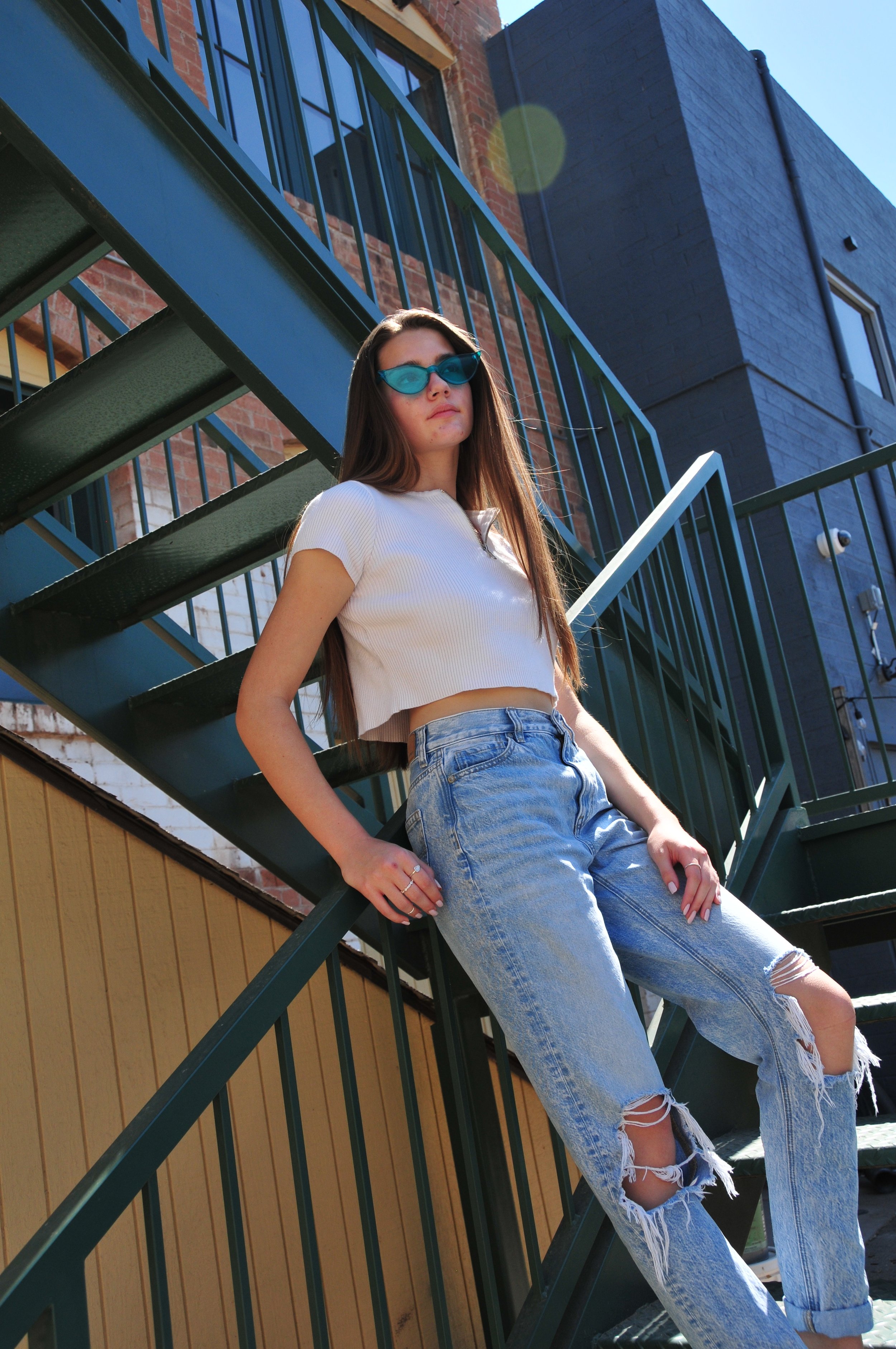 A young woman with long brown hair, wearing blue sunglasses, a white crop top, and ripped jeans, is leaning against a metal staircase outdoors on a sunny day.