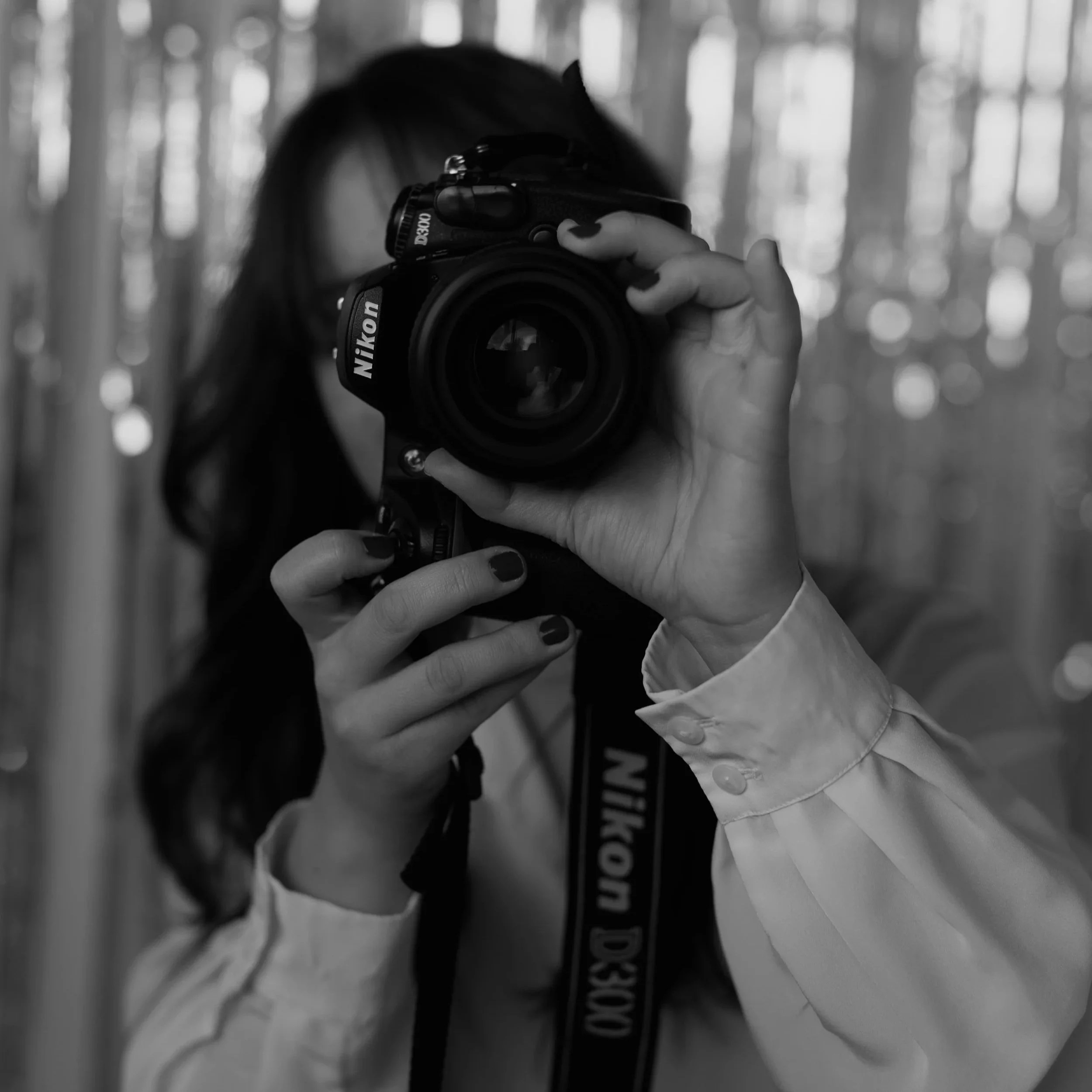 Black and white photo of a person taking a self-portrait with a Nikon camera, reflected in a mirror, with blurred background of trees or vertical lines.