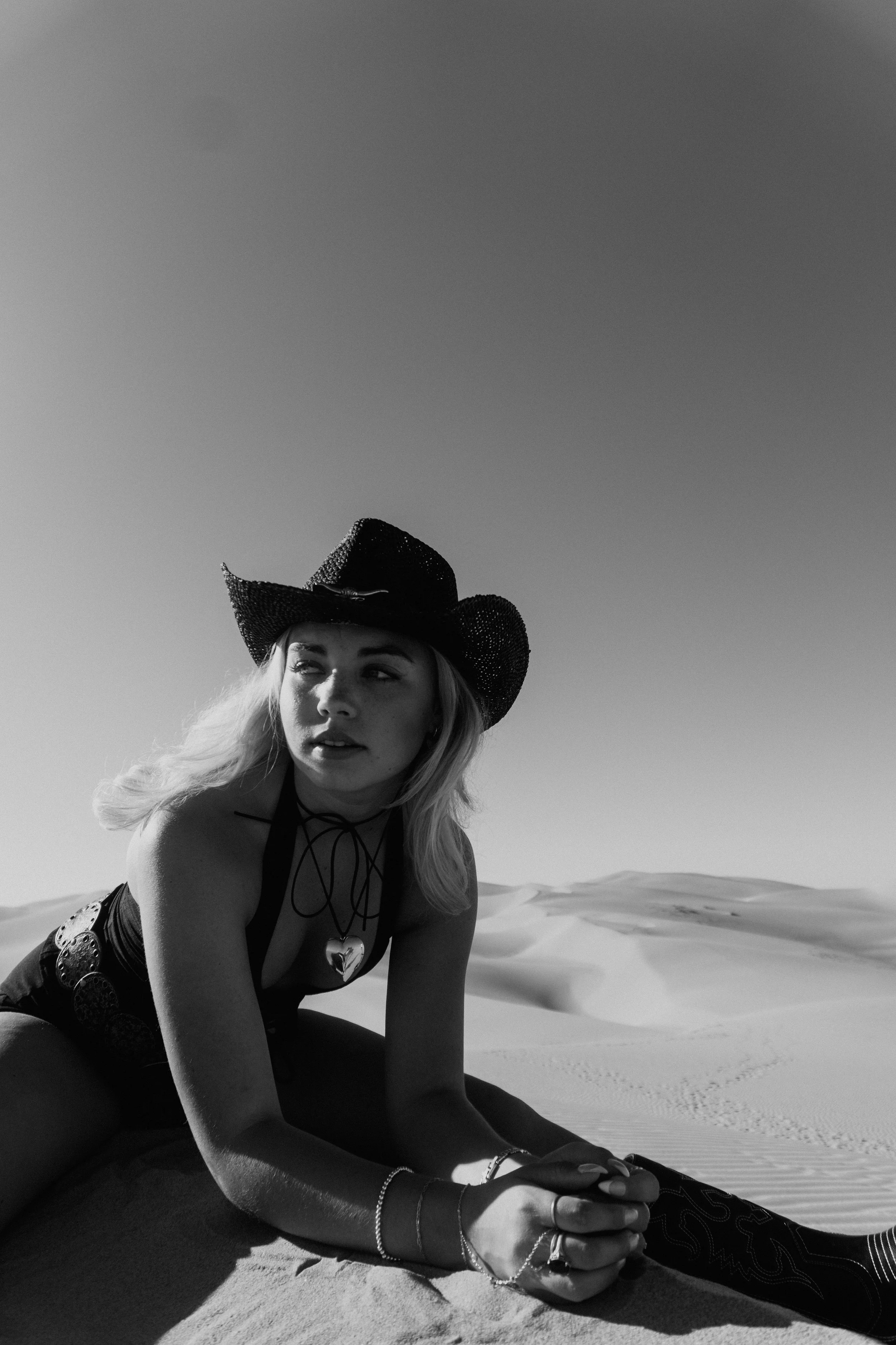 A woman with long hair wearing a cowboy hat and jewelry sitting on sand dunes in a desert landscape.