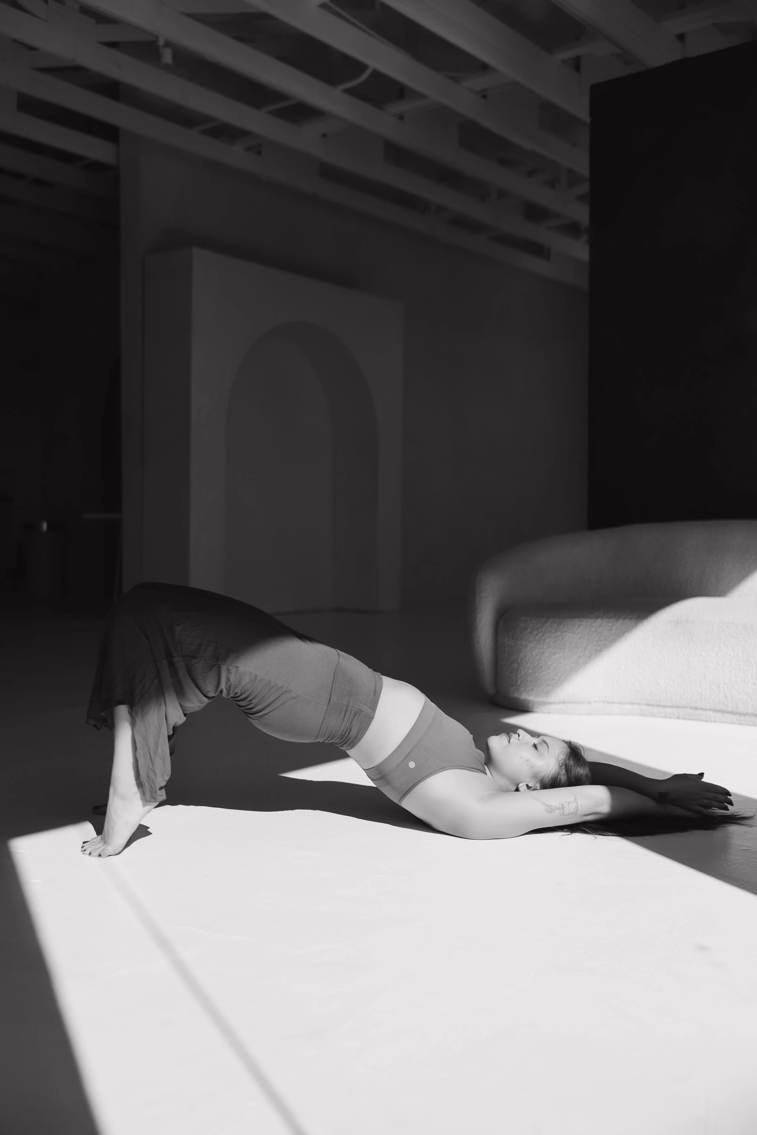 A woman practicing yoga indoors, performing a pose with her legs bent backward and her arms extended overhead, lying on a mat with furniture and a wall in the background.