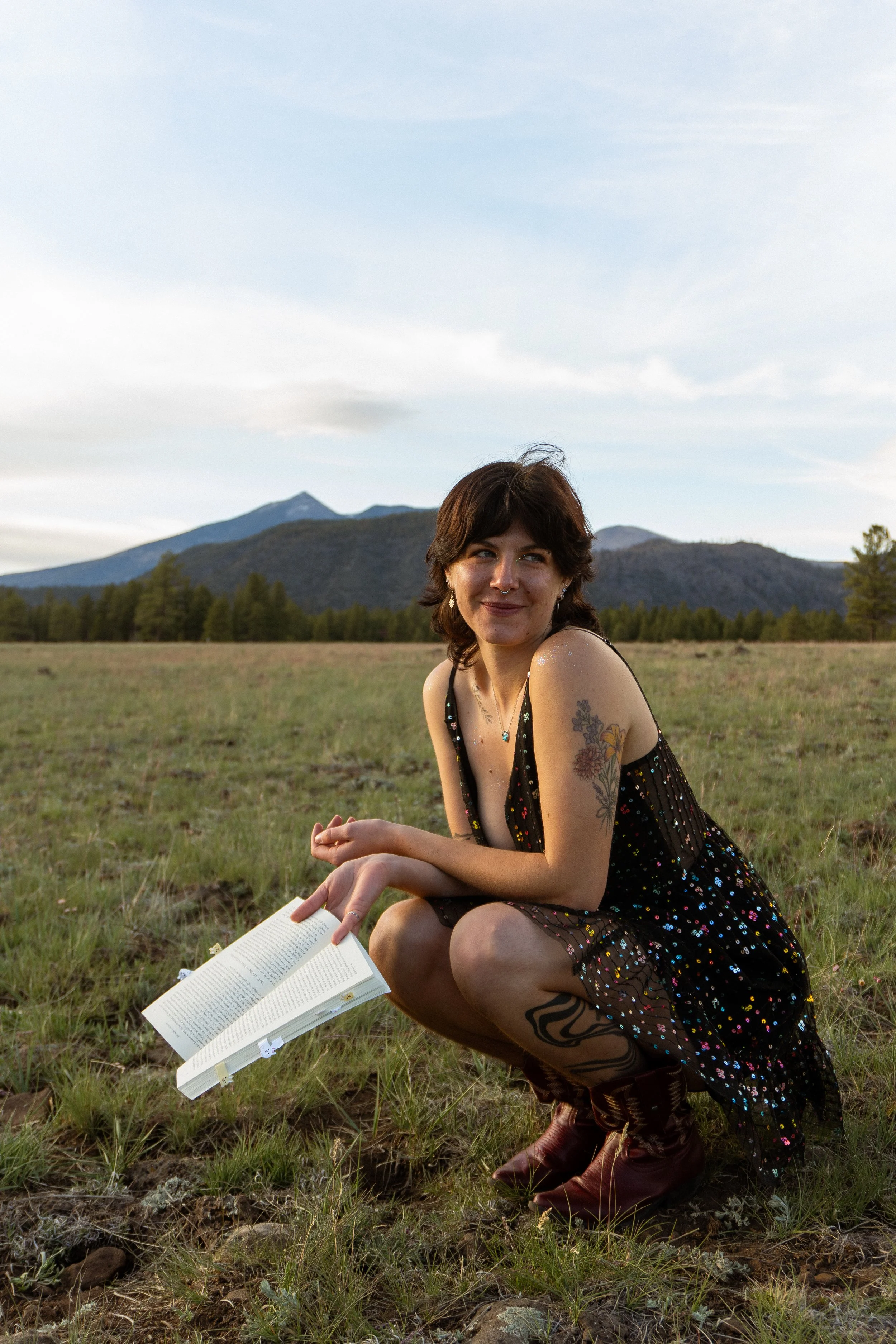 Young woman with short brown hair, tattoos, and septum piercing, wearing a black sequined dress, squatting in a grassy field with mountains in the background, holding an open book.