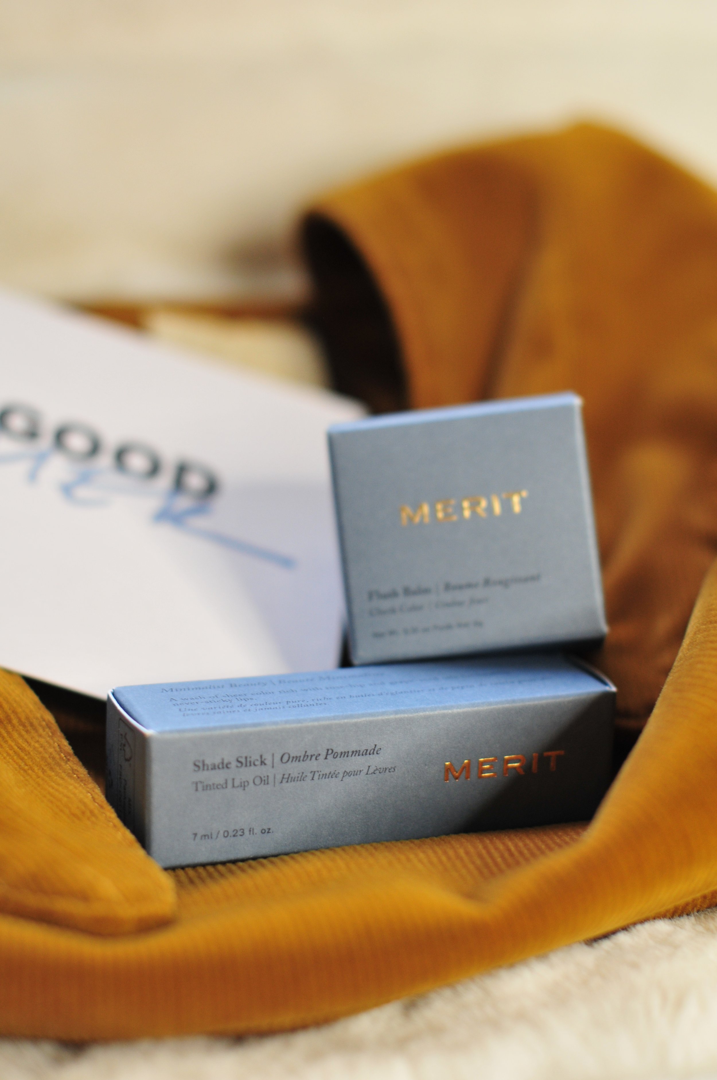 Close-up of two small skincare product boxes labeled 'MERIT,' placed on a soft surface with a mustard-yellow fabric surrounding them.