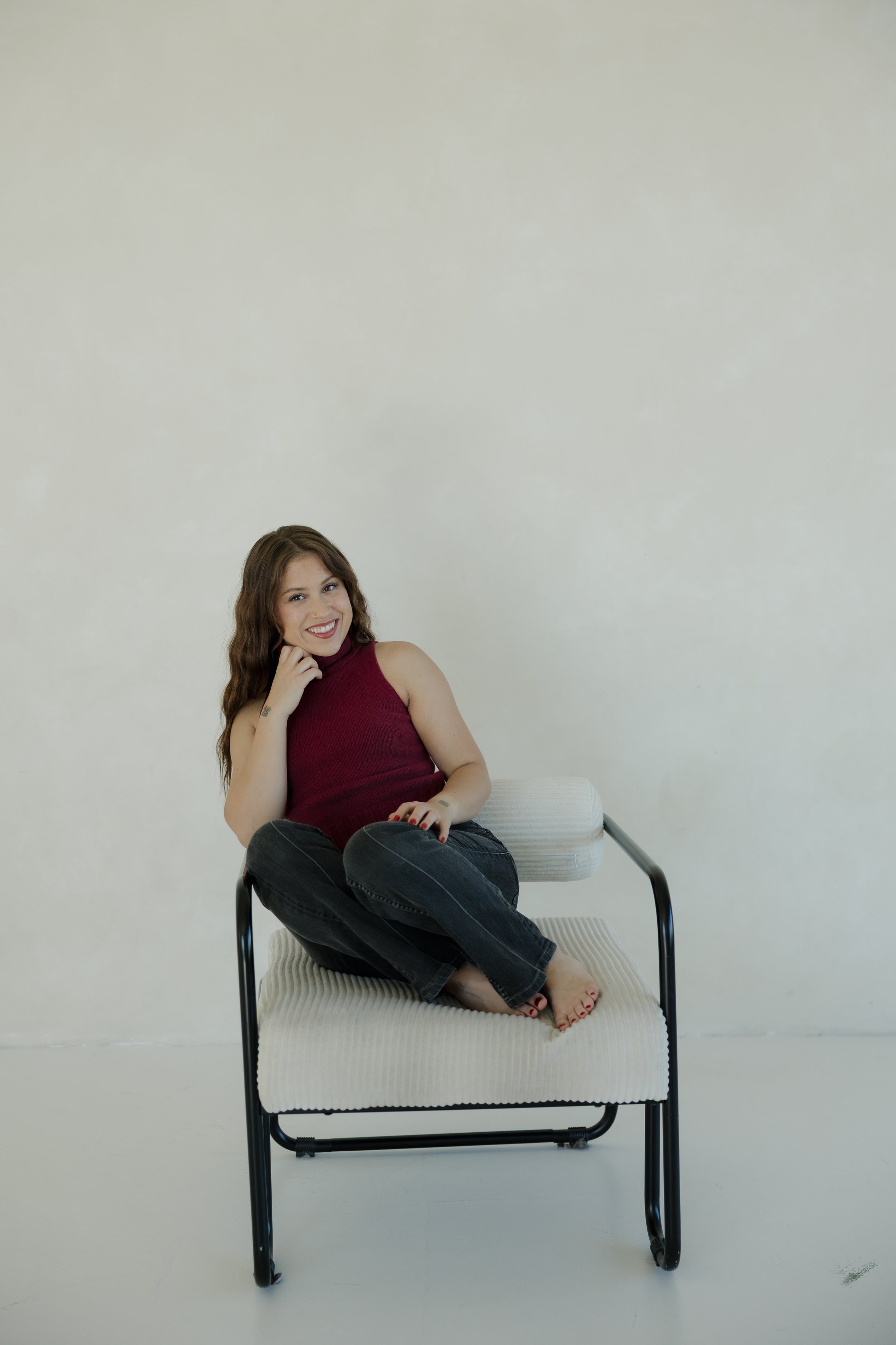 A young woman with long wavy brown hair smiling and sitting cross-legged on a white chair, wearing a sleeveless maroon top and black jeans, against a plain off-white background.