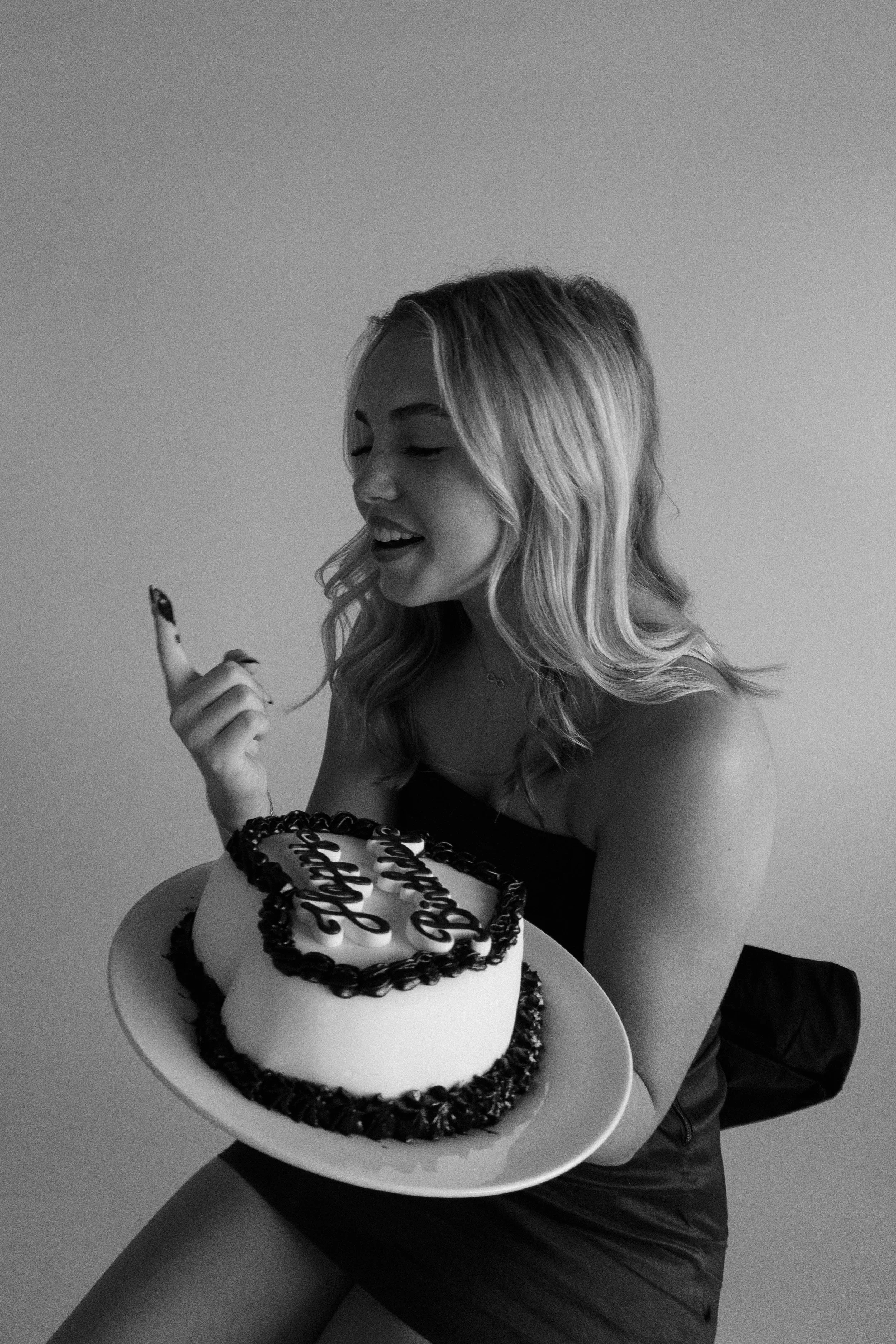 A young woman with wavy hair holding a birthday cake with the words 'Happy Birthday' written on it, smiling and looking at the cake.