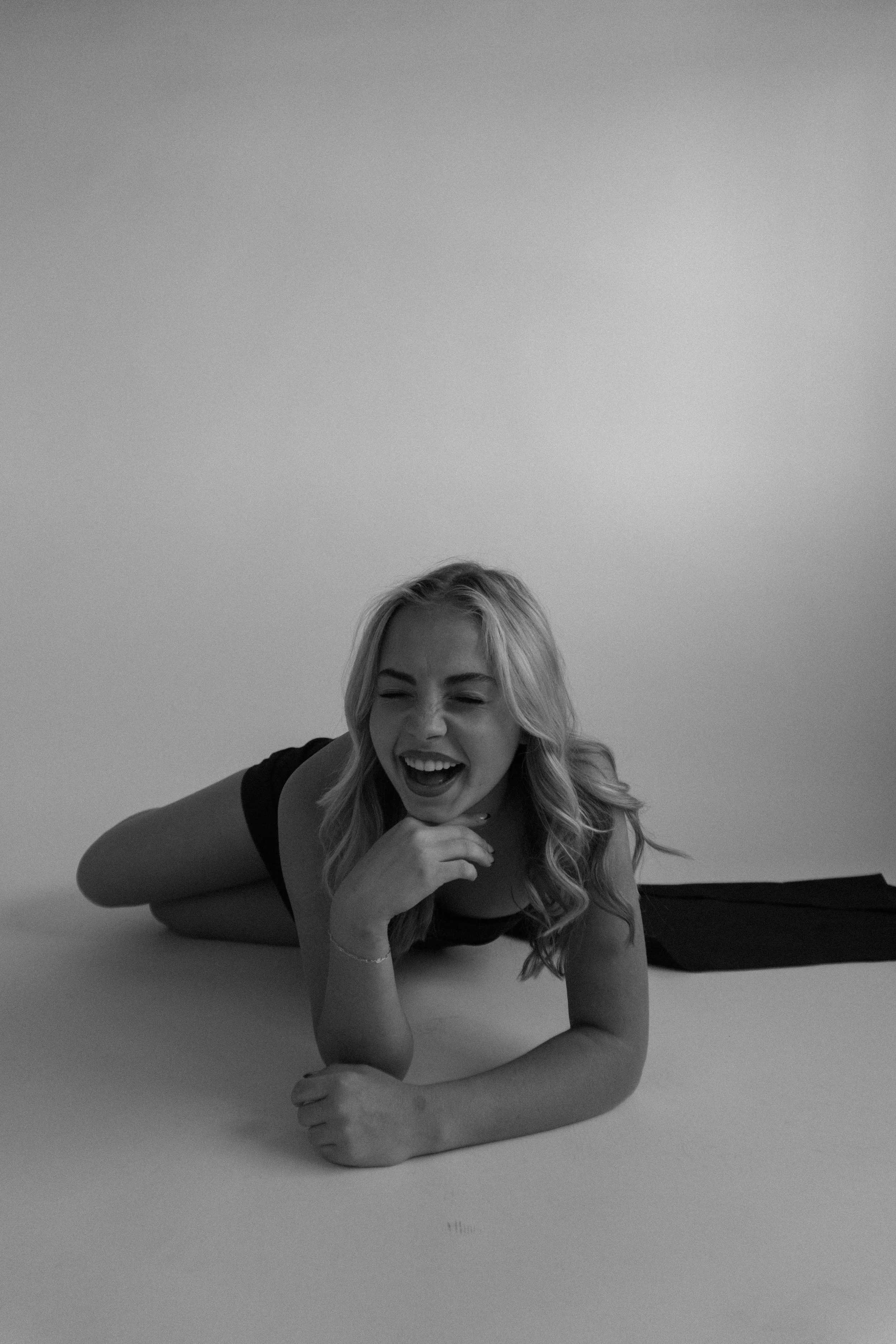 A woman with long blond hair lying on her side on a white surface, smiling and laughing with her hand near her chin. She is wearing a dark sleeveless top and jeans.