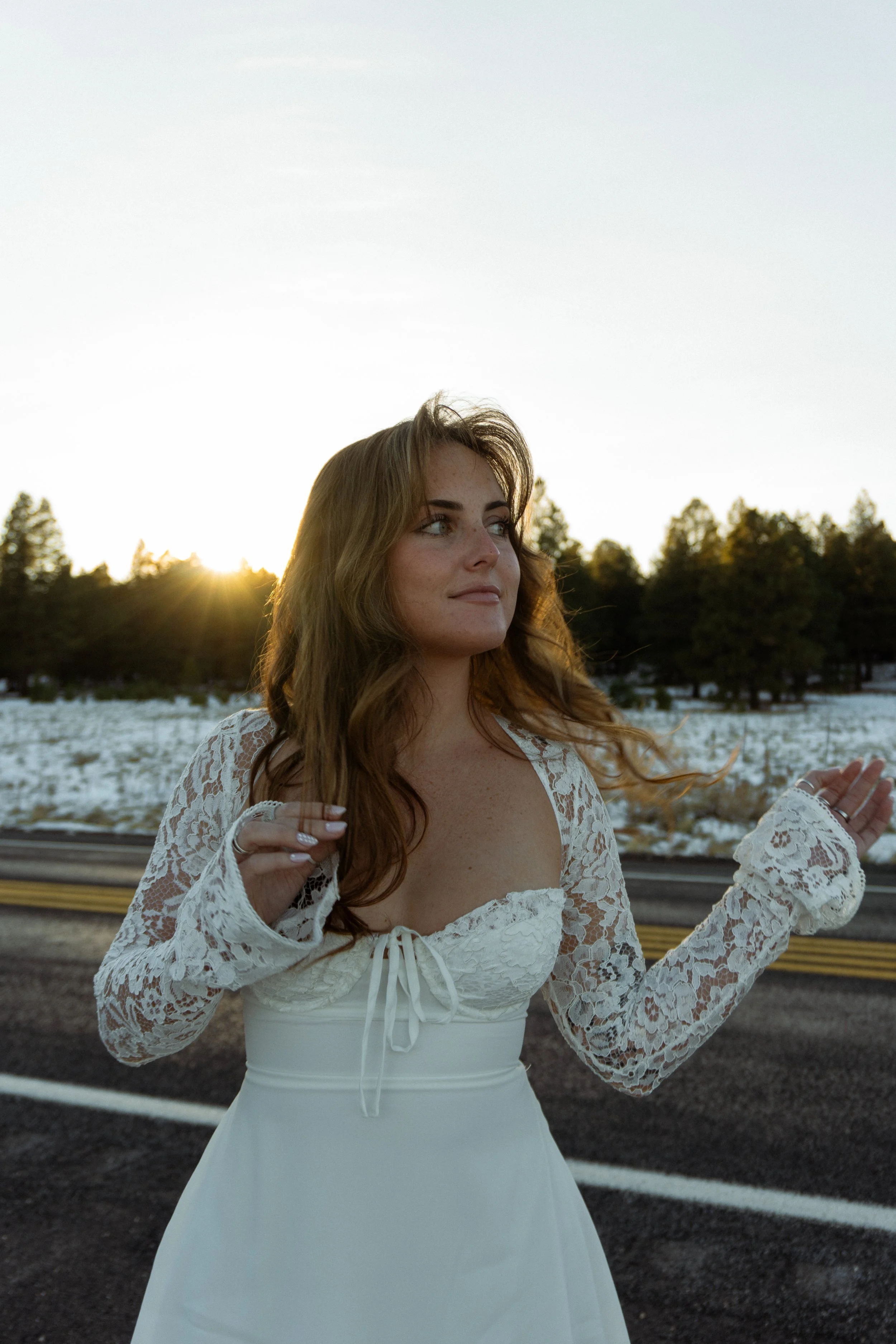 A woman with long, wavy, brown hair standing outdoors at sunset, wearing a white dress with lace long sleeves and a lace-up corset-style bodice, in front of a wooded area and a fence.
