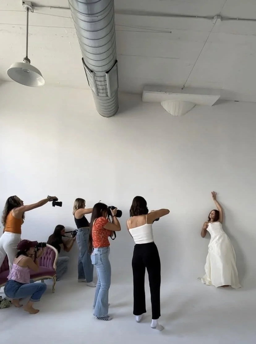 A woman in a white dress is posing against a white studio wall with her arm raised. Six women, some with cameras, are taking her photos in a studio setting with studio equipment visible on the ceiling.