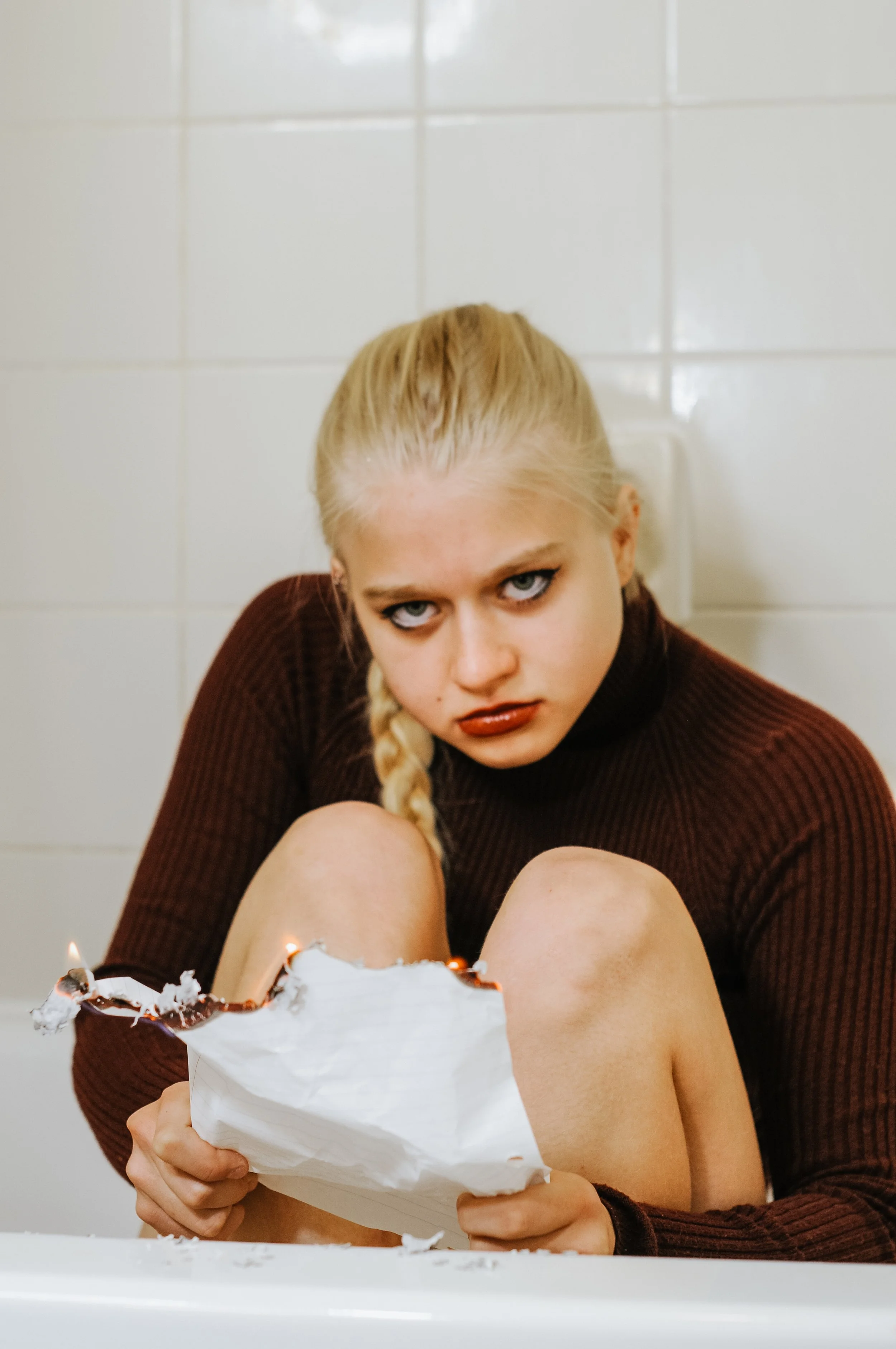 A woman with platinum blonde hair in a braid, wearing a brown sweater, is sitting in front of a white tiled wall, holding a white paper bag with a broken cake inside, with some candles and frosting visible.