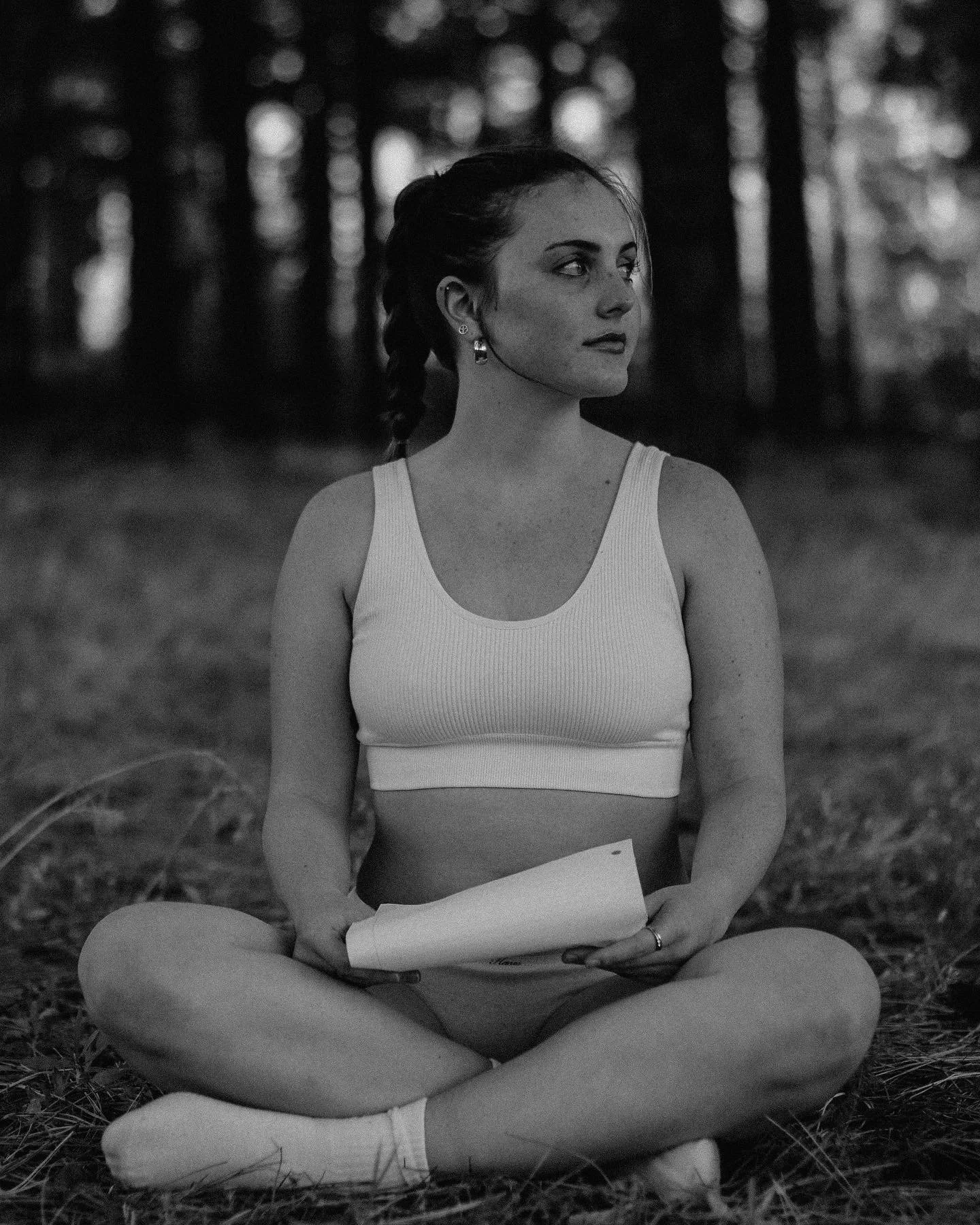 A woman with dark hair in a braid, wearing earrings, a sports bra, and leggings, sitting cross-legged outdoors holding papers, with a blurred forest background in black and white.