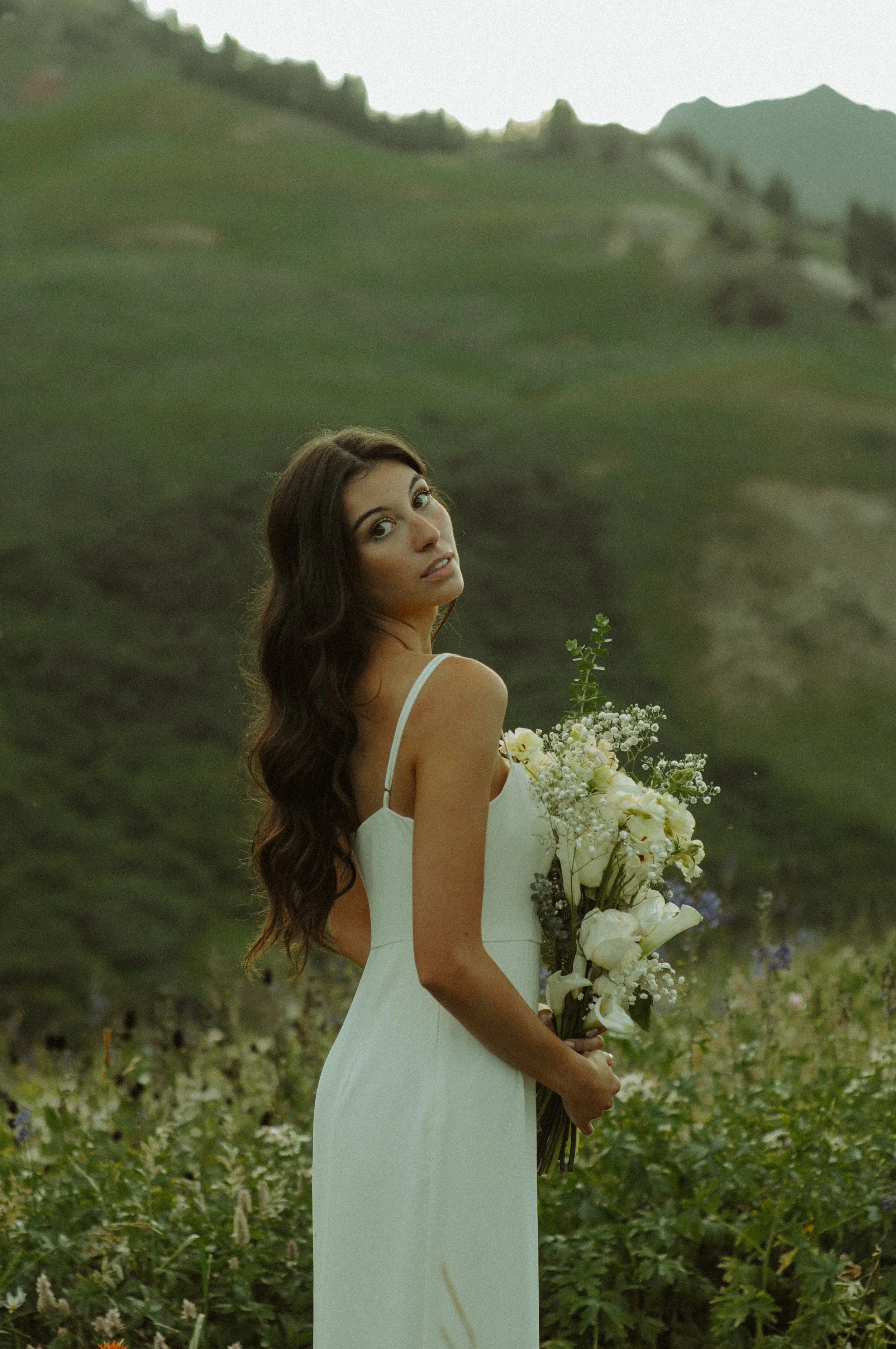 A woman in a white dress holding a bouquet of white flowers in a field of greenery, with mountains in the background.