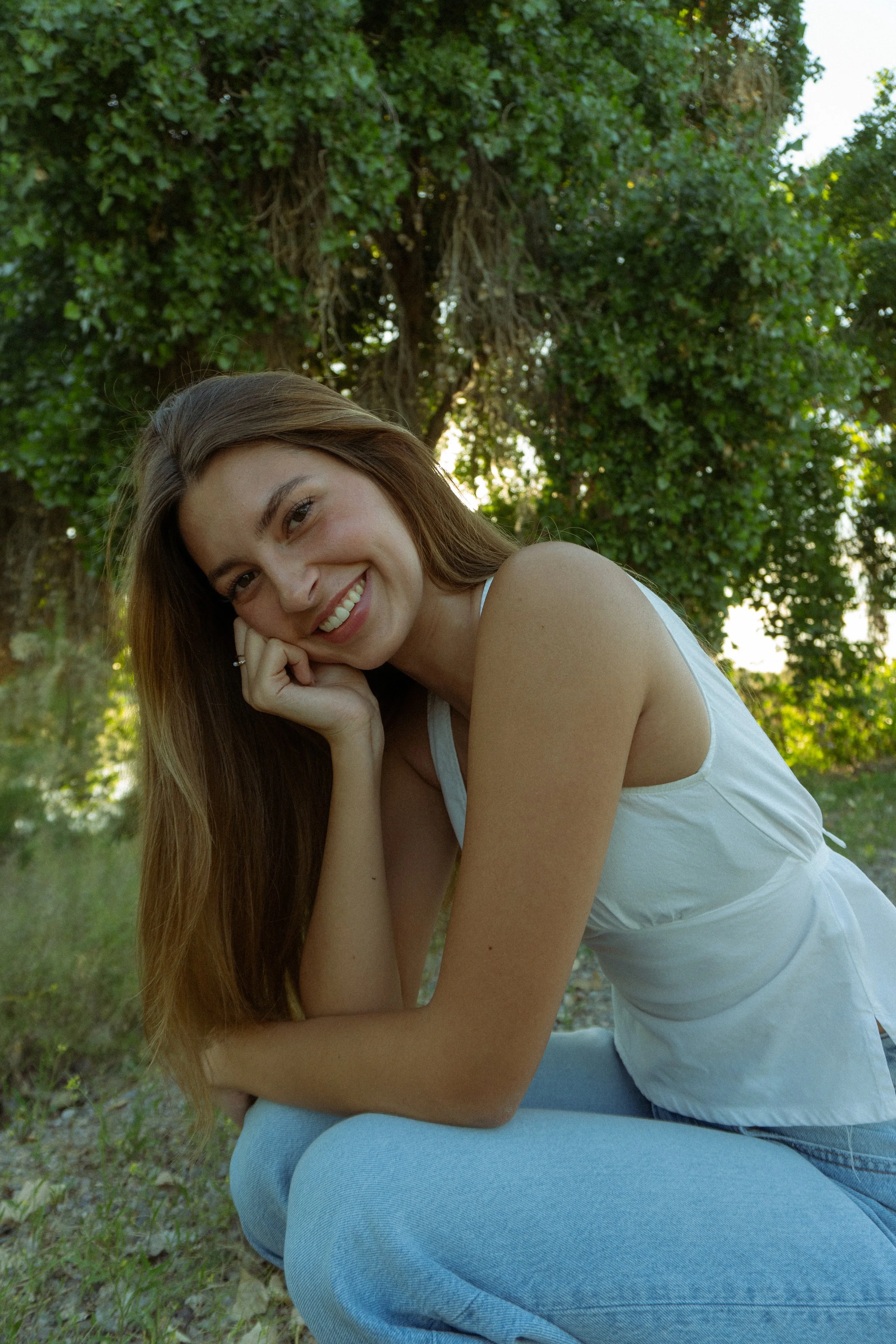A young woman with long brown hair smiling while sitting outdoors on grass, wearing a white tank top and light blue jeans, with a large leafy tree in the background.