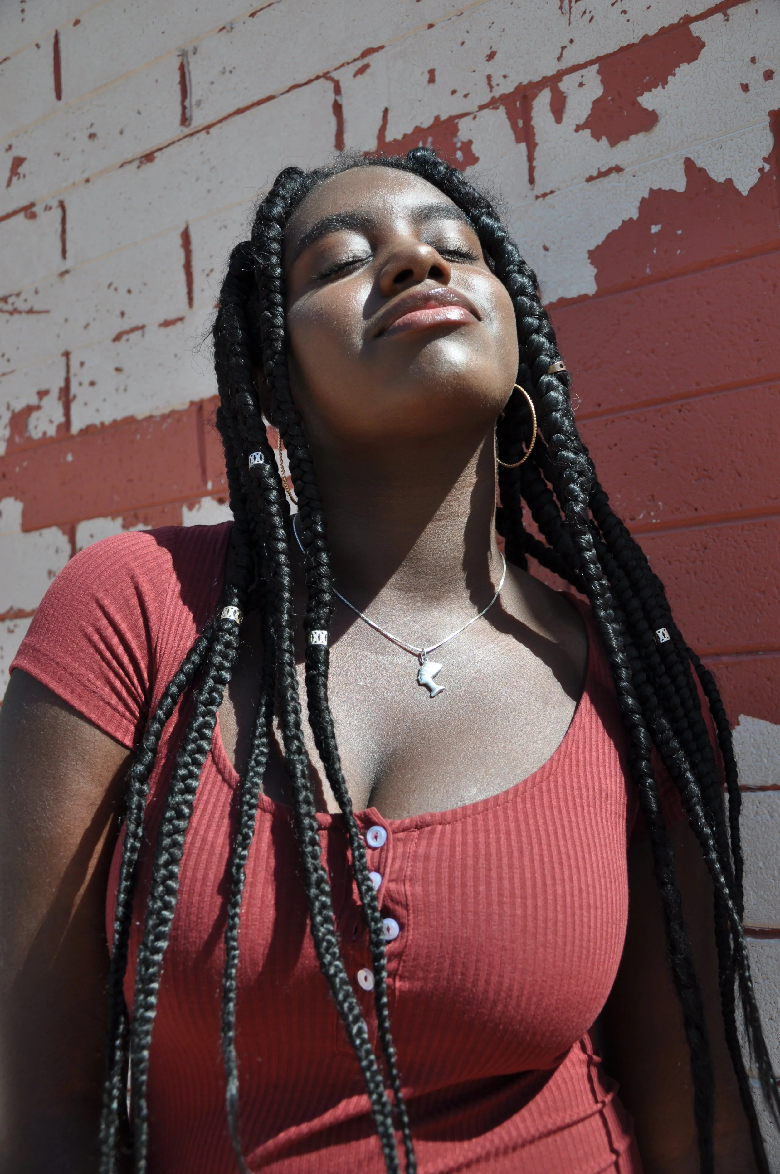 A woman with long braids, wearing hoop earrings, a necklace with Africa shaped pendant, and a red button-up shirt, leaning against a painted brick wall with her eyes closed.