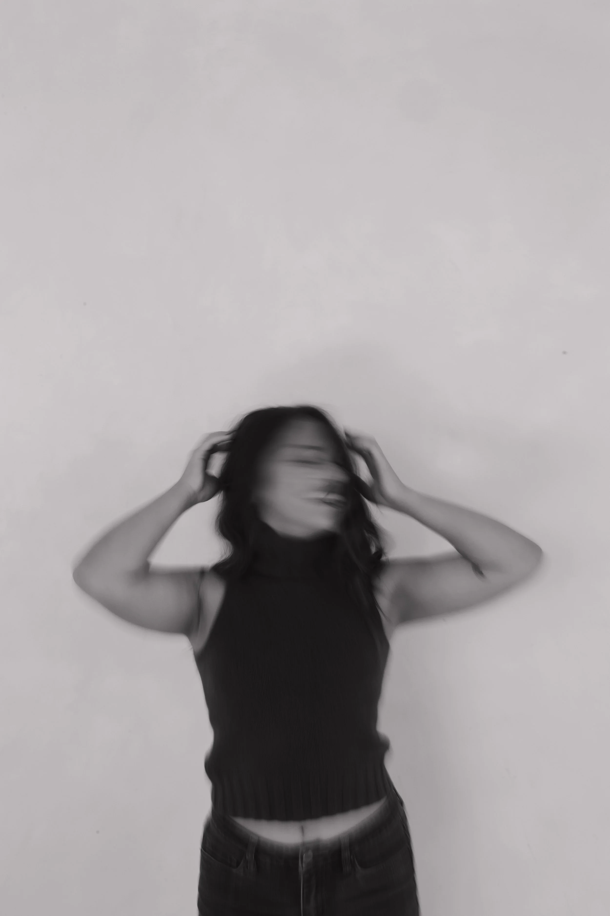Black and white photo of a woman adjusting her hair, wearing a sleeveless top and jeans, standing against a plain wall.