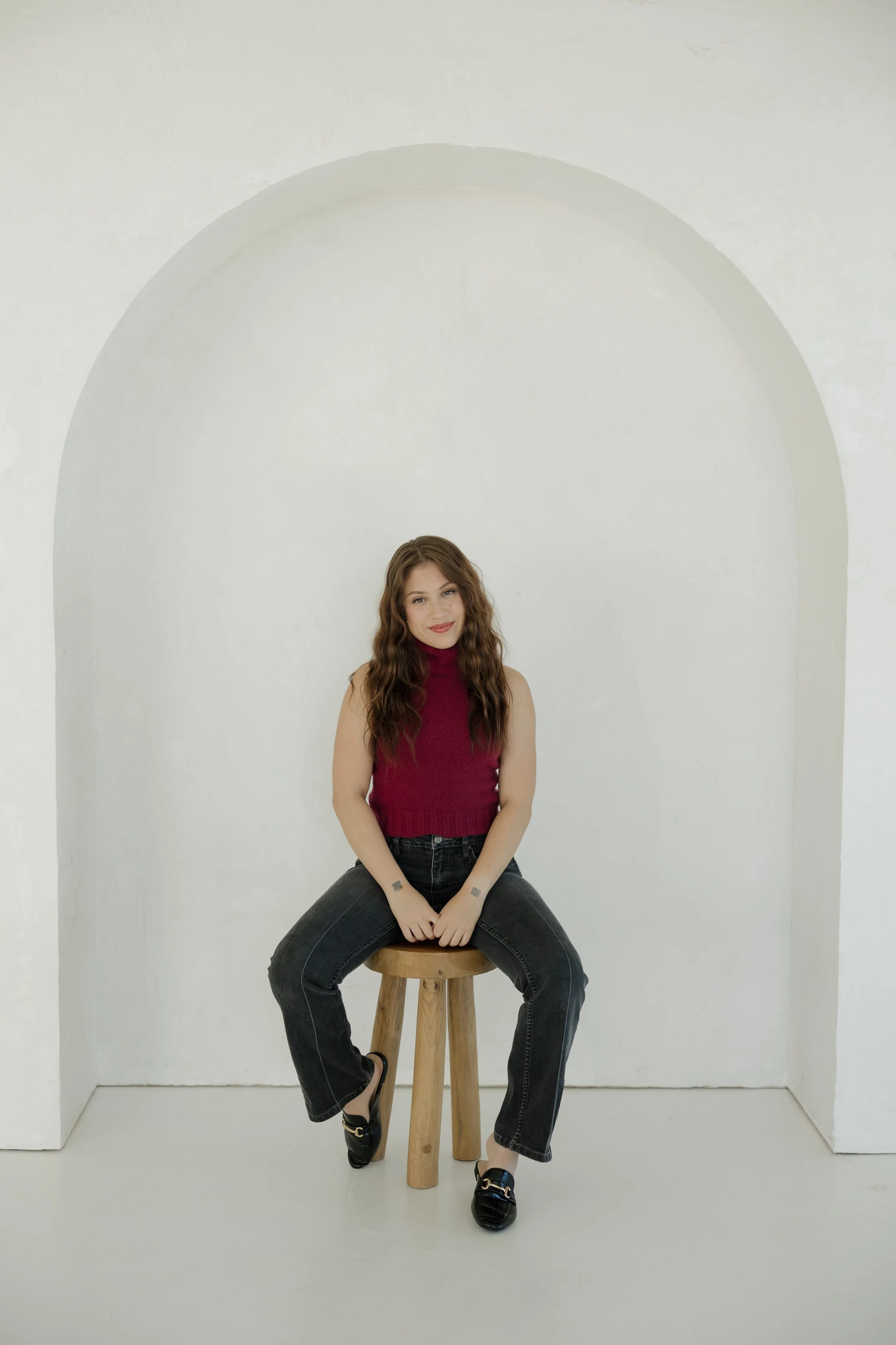 A young woman with long, wavy brown hair and a tattoo on her wrist, sitting on a stool with her legs apart. She is wearing a sleeveless maroon top, black jeans, and black slip-on shoes with gold accents. She is smiling, with her hands resting on her thighs, in a minimalist white room with an arched wall behind her.