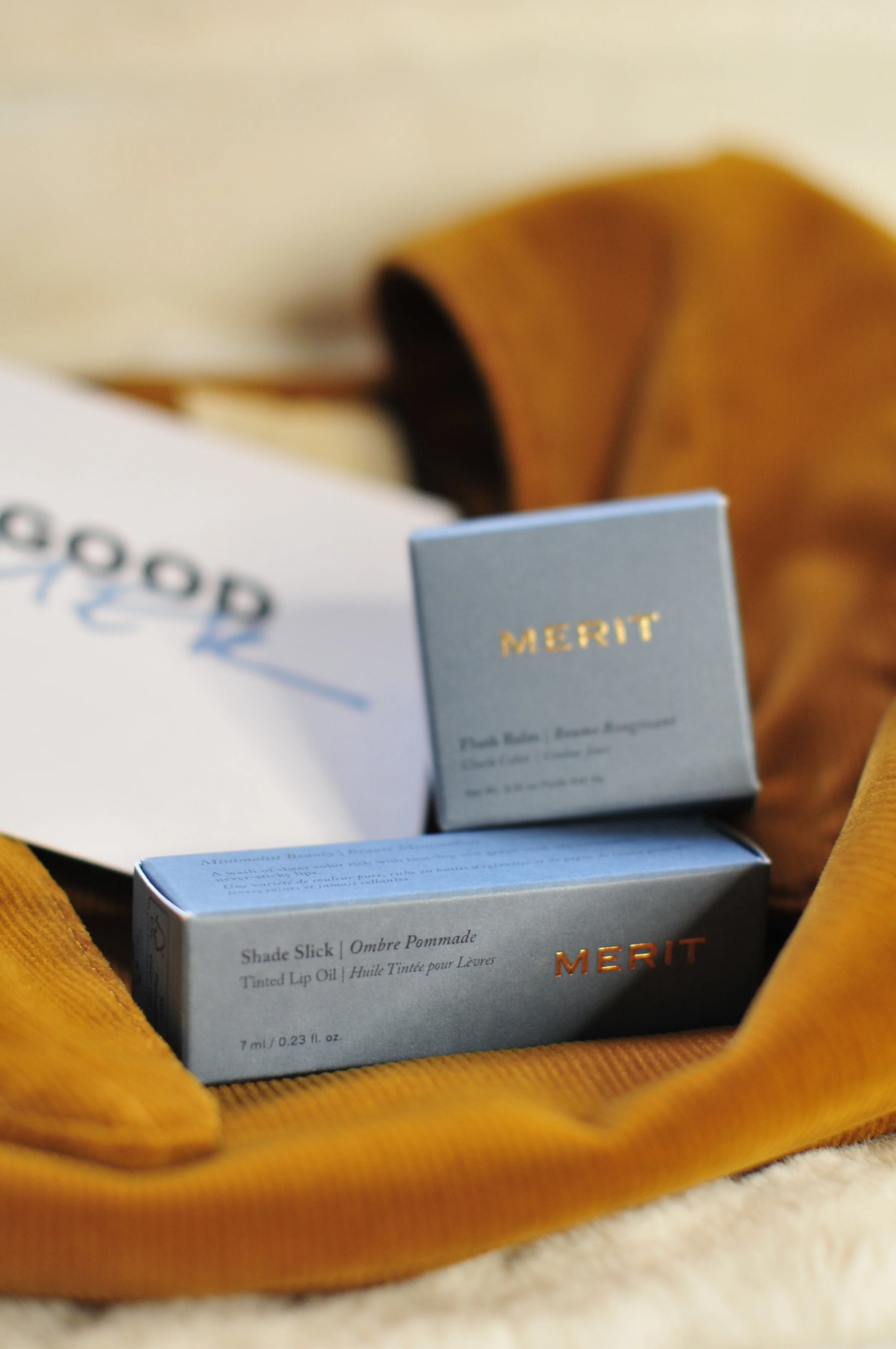 Close-up of blue and white Merit skincare and makeup product boxes, labeled 'Shade Slick' and 'Ombre Pommade,' inside a soft, orange, textured bag on a beige surface.