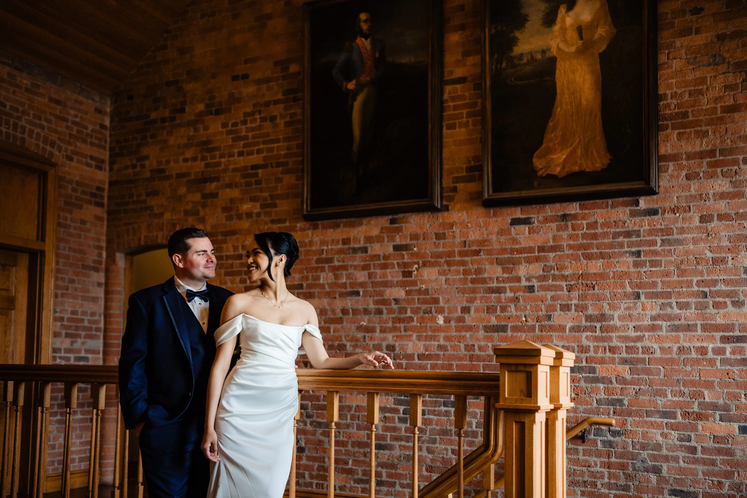 A wedding couple standing on a staircase with a brick wall background and framed paintings hanging above them.