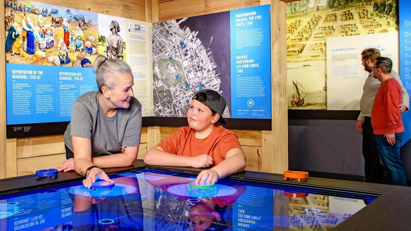 A woman and boy playing air hockey at a museum exhibit, with colorful informational panels on the wall behind them.