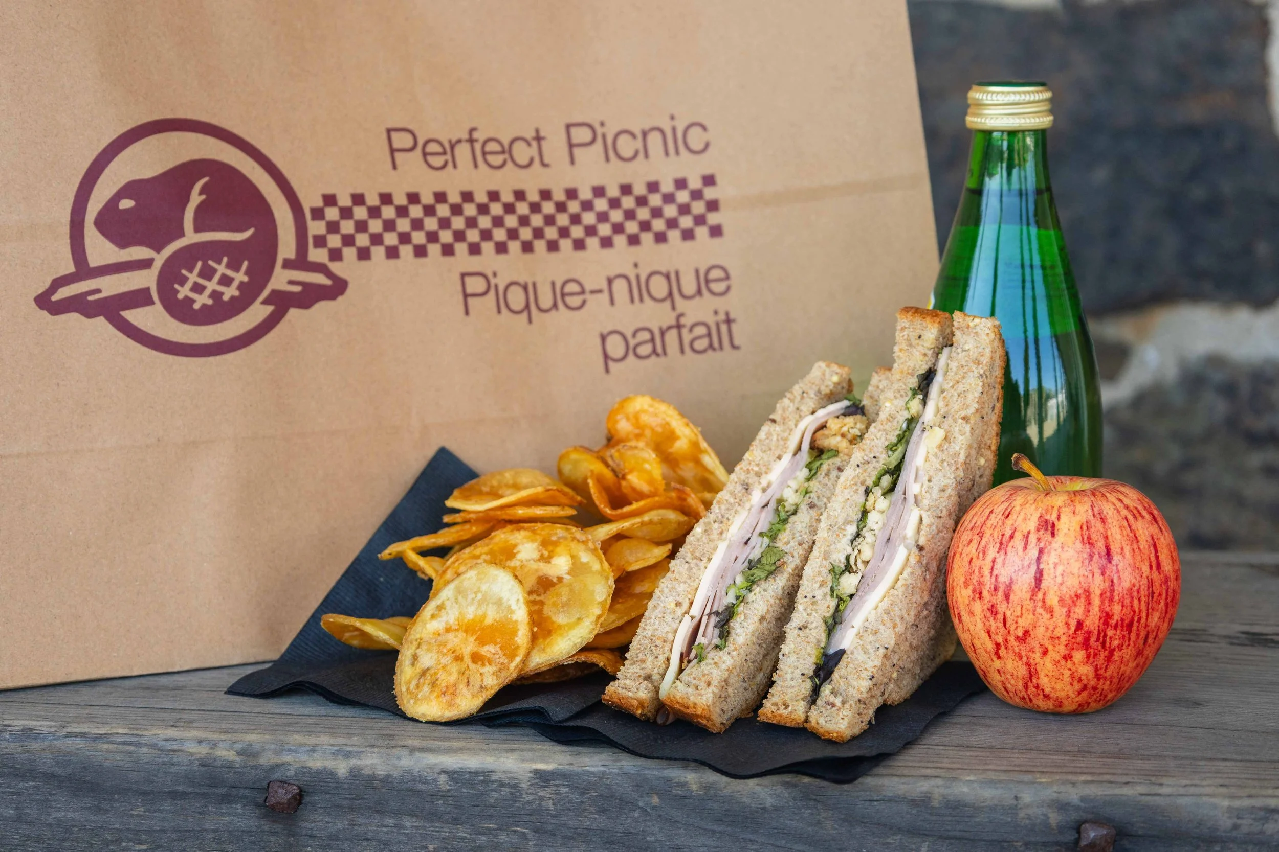 Picnic meal with a wrapped sandwich, potato chips, a red apple, a green bottled beverage, and a paper bag with a logo and text in French and English.