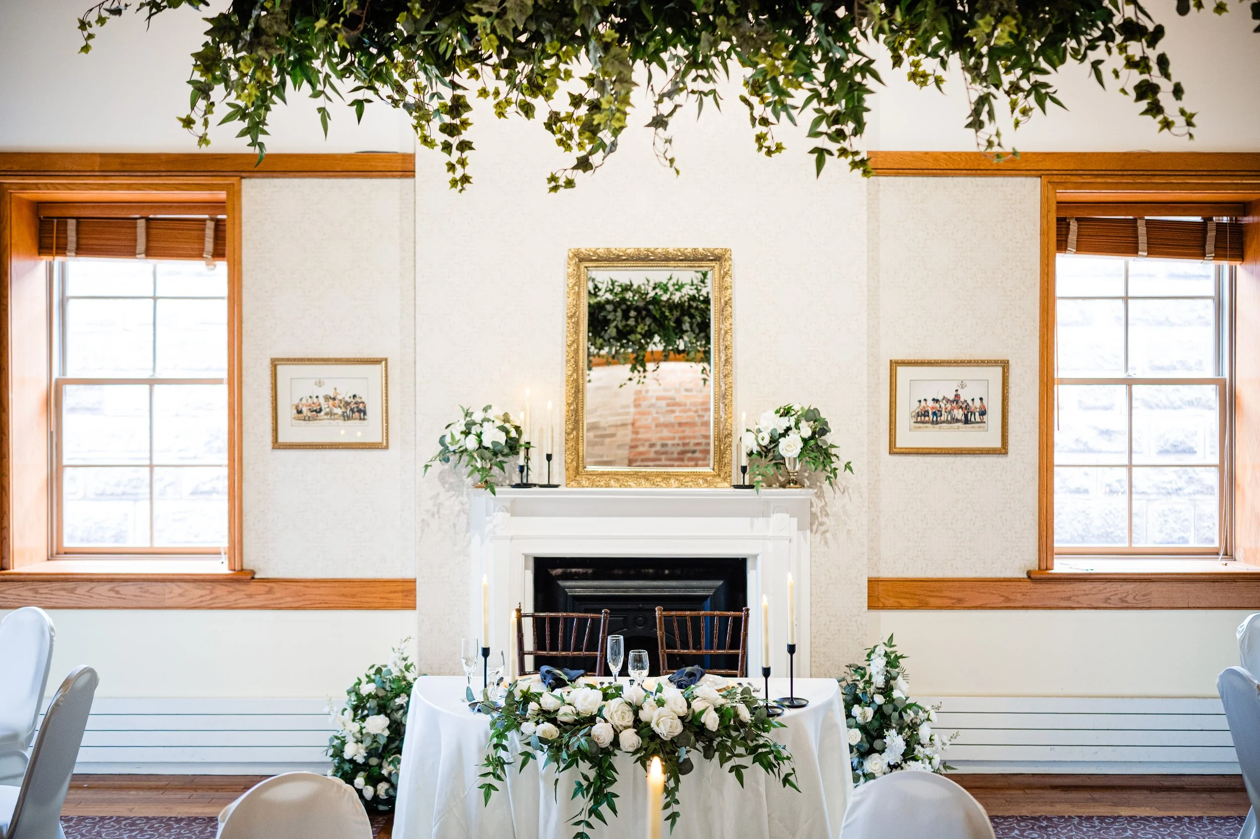 Elegant dining room setup with a white tablecloth-draped table decorated with white flowers and greenery, surrounded by white chairs, a fireplace with a large framed mirror and white floral arrangements, two large windows with wooden trim, and green ivy hanging from the ceiling.