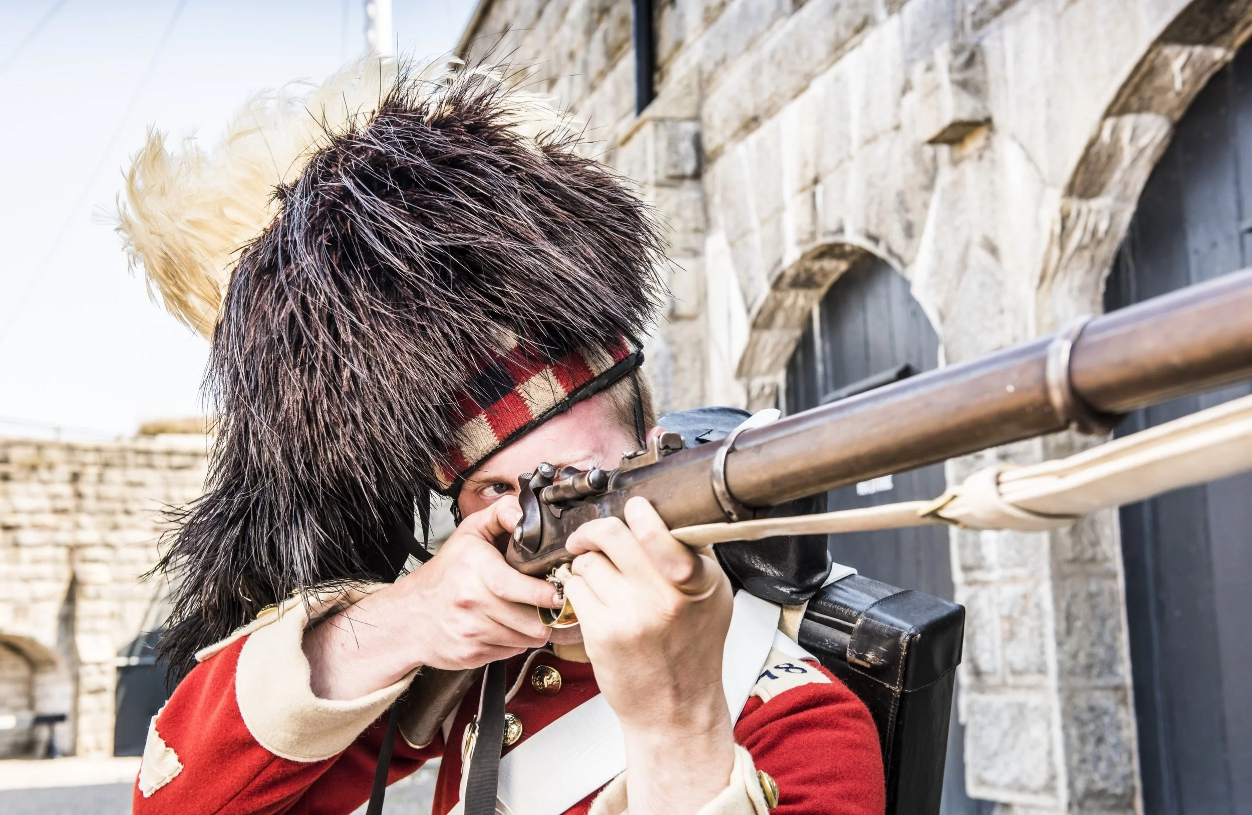 Person dressed as a historical soldier, wearing a fur hat and red military uniform, aiming a long rifle.