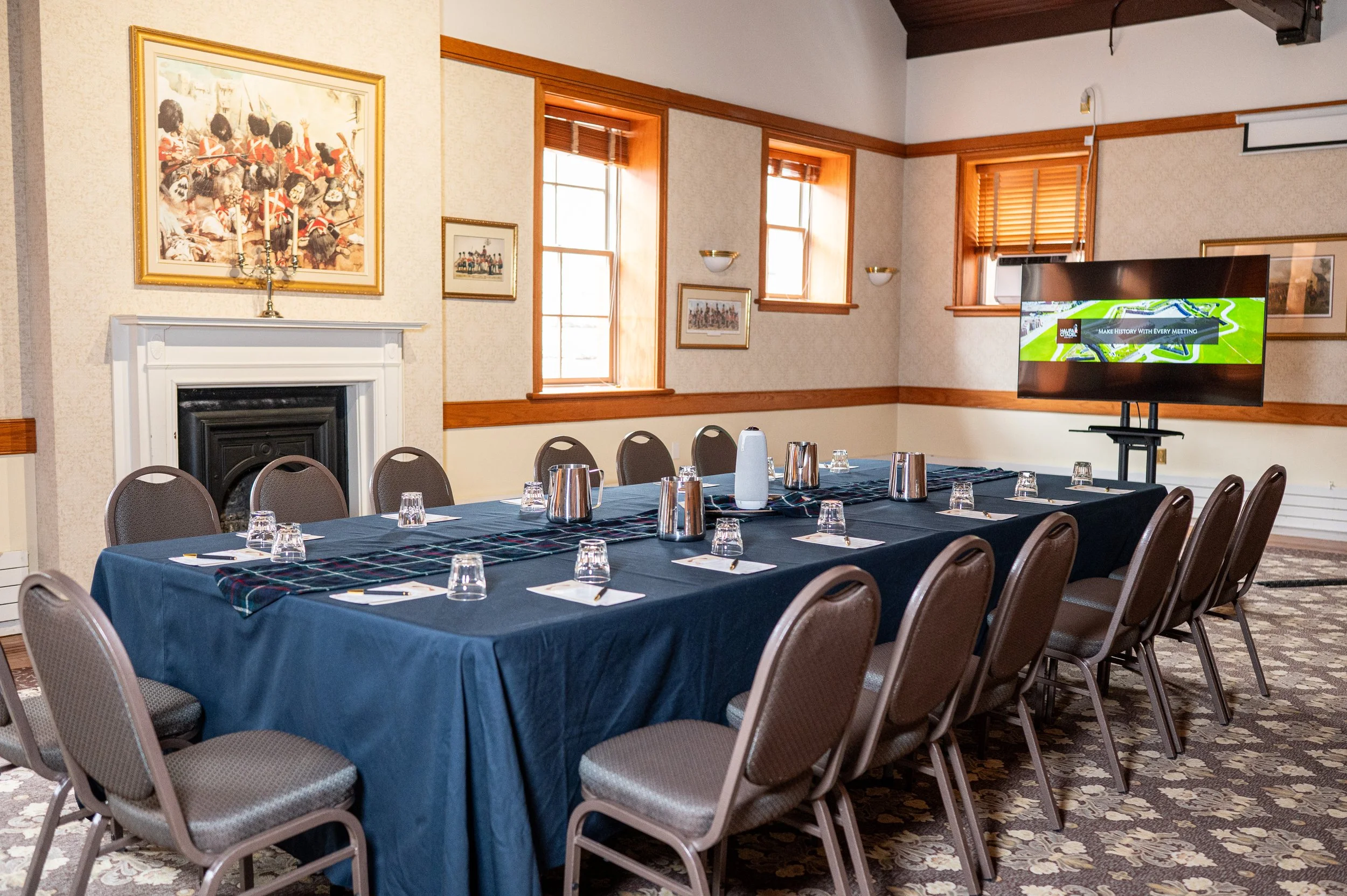 A conference room with a long table covered with a dark blue tablecloth, surrounded by chairs. The table is set with glass cups, metal pitchers, and plates. There are five windows with wooden blinds on the wall, which let in natural light. A large flat-screen TV is positioned on a stand, displaying a graphic with green and black colors. The walls have framed artwork, including a prominent painting of soldiers in red uniforms. A decorative fireplace with a white mantel is visible beneath the painting.
