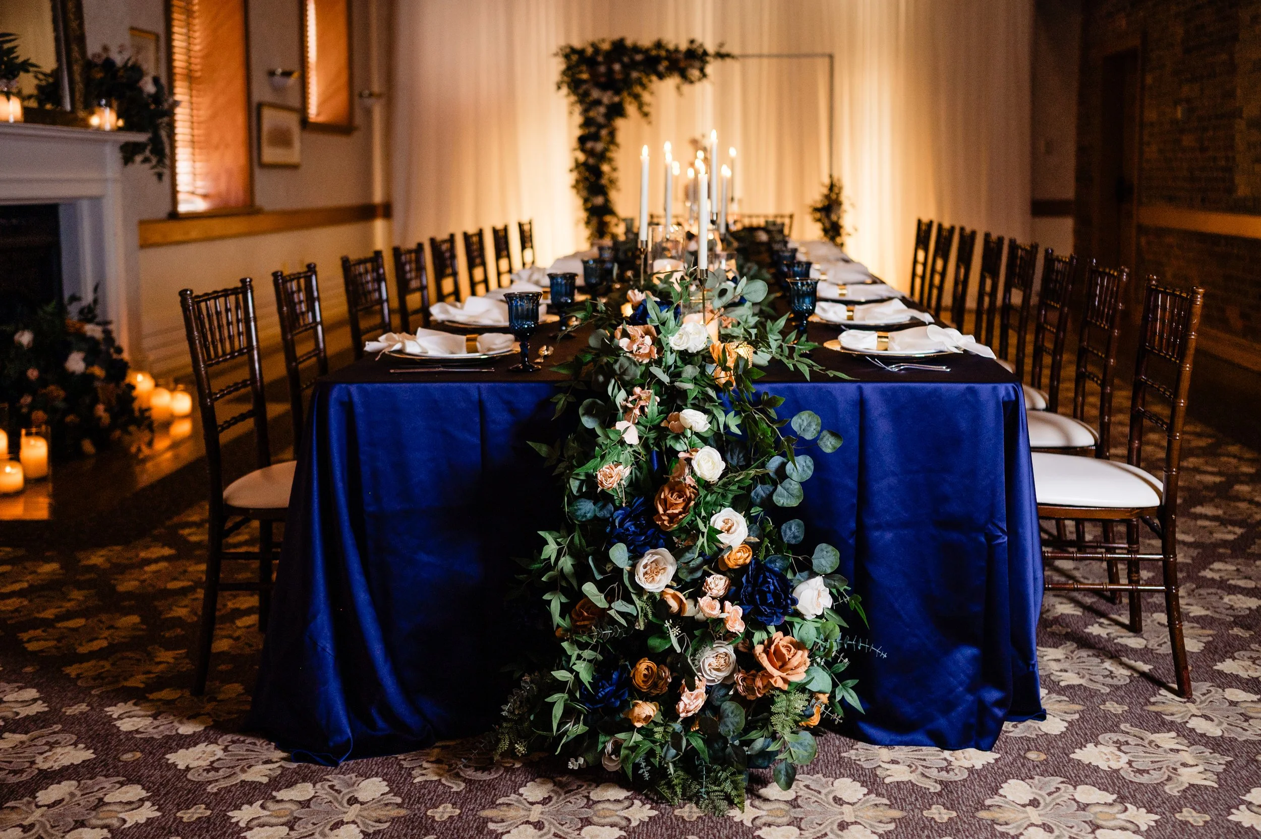 A long banquet table decorated with a lush floral runner and tall white candles in a dimly lit room for a celebration or wedding.