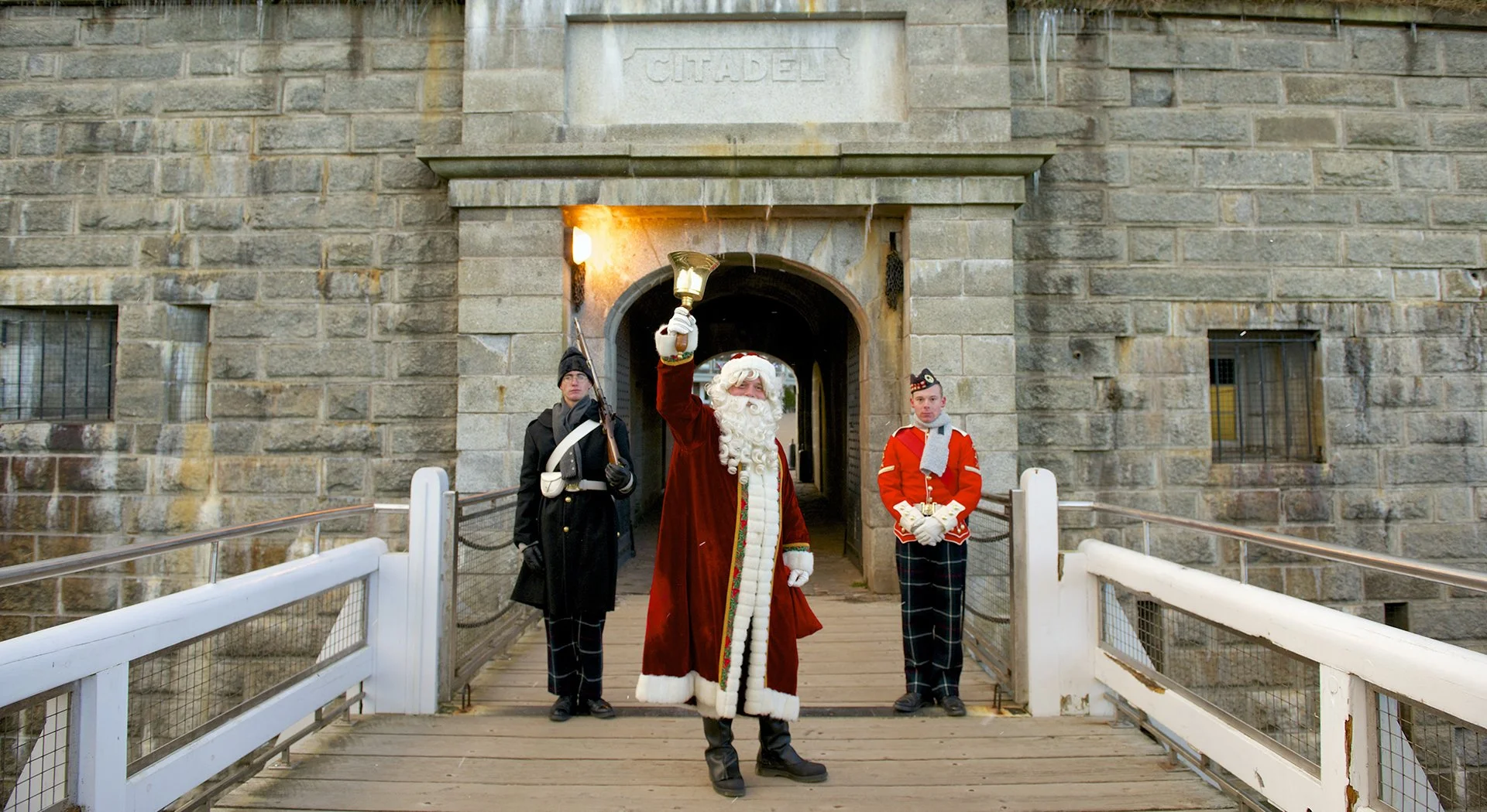Santa Claus holding a bell and waving, flanked by two men in military uniforms, at a historic stone fort entrance with a sign labeled Citadel above.