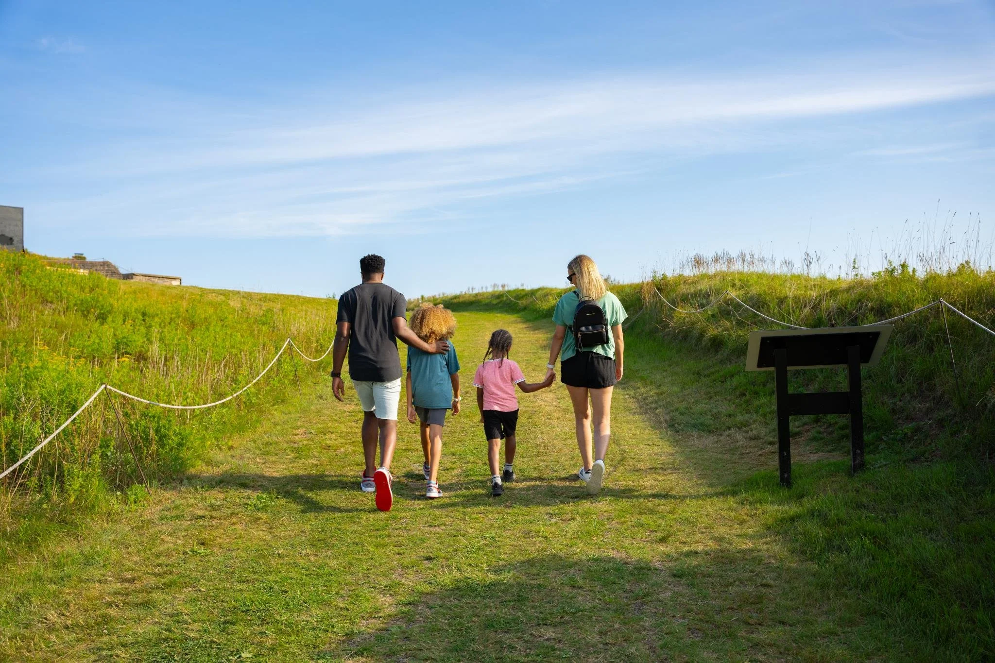 A family walking along a grassy path in a park under a blue sky. The group includes a man, woman, and two children, some holding hands, with a sign and informational plaque nearby.