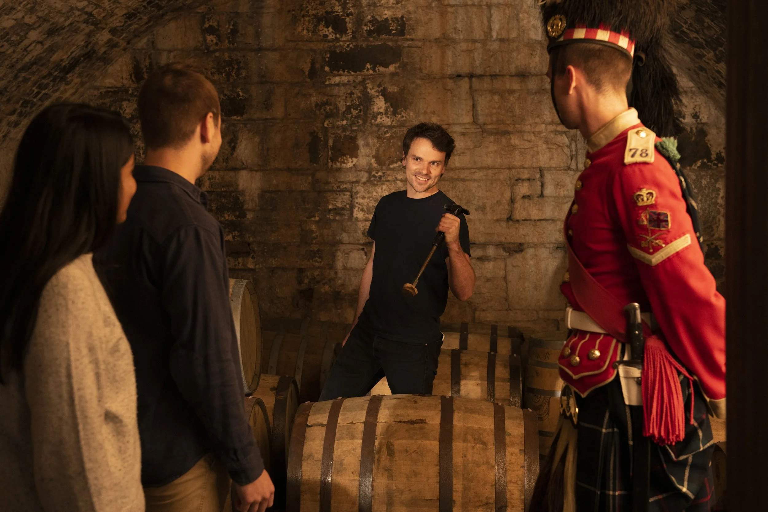 Group of three people in a wine cellar, one person dressed as a 78th Highlander, the others in casual clothing, standing near a large wine barrel and stone walls.