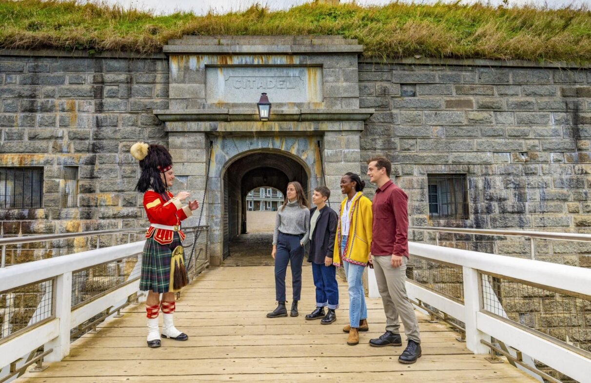 A group of four people standing on a bridge outside the Citadel, listening to a person dressed in 78th Highlander uniform who appears to be giving a tour or explanation.
