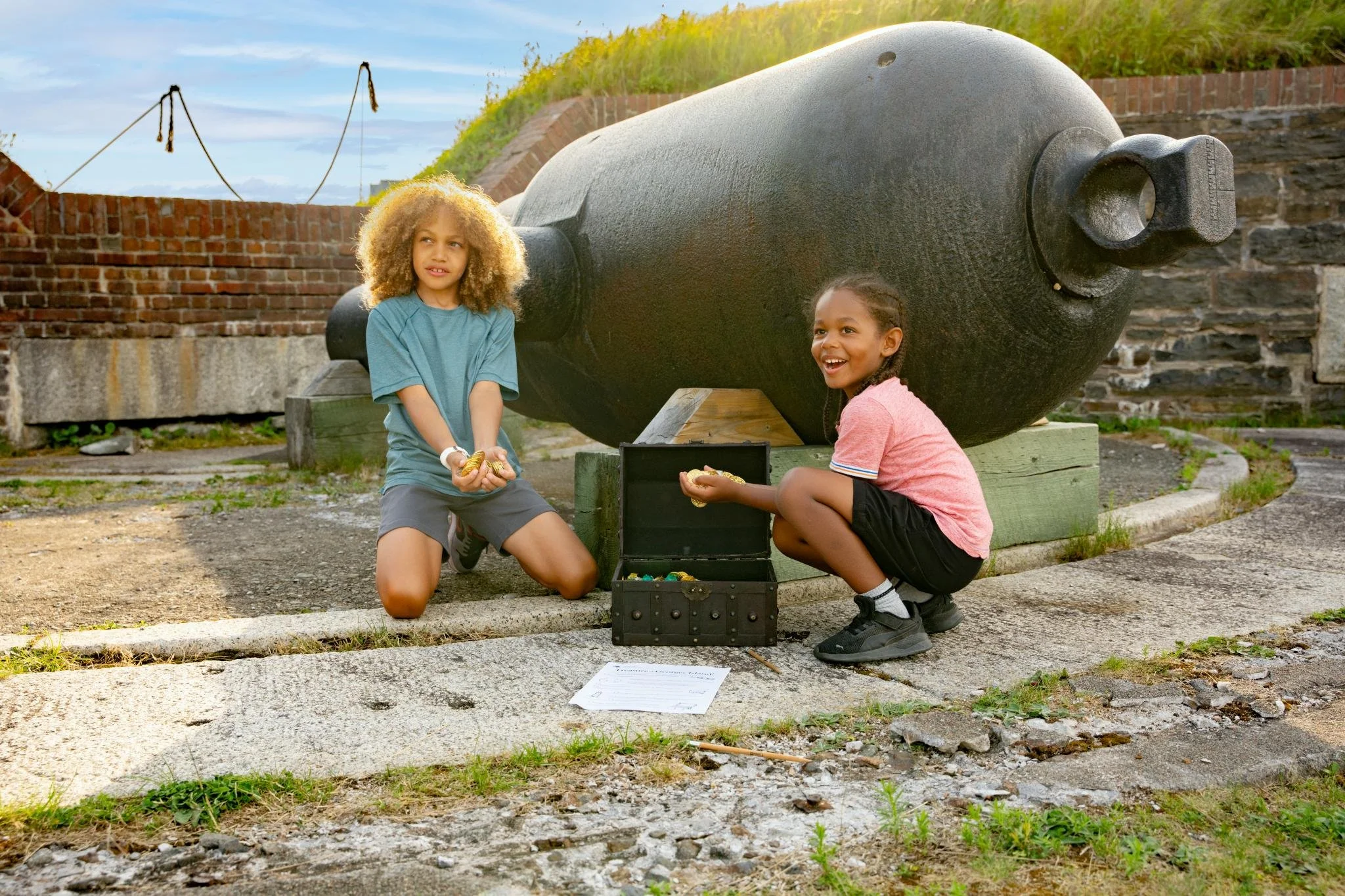 Two children kneeling on the ground next to a large cannon, with open treasure chest filled with gold coins between them, in an outdoor setting with brick wall and grassy hill in the background.