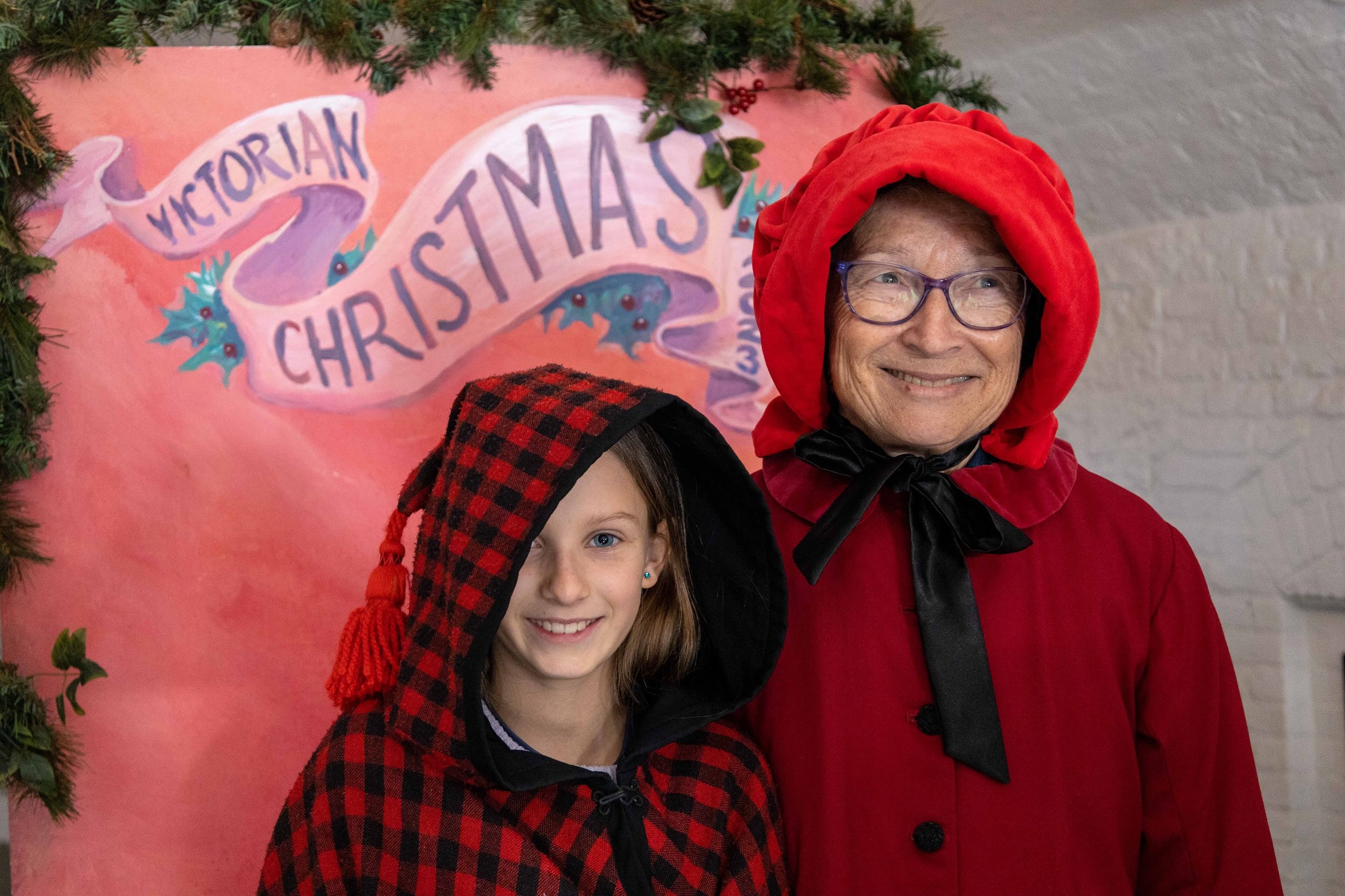 A woman wearing a red hooded robe and glasses standing next to a young girl in a red and black checkered hooded jacket, both smiling in front of a pink Christmas sign that reads 'Victorian Christmas' decorated with green foliage.