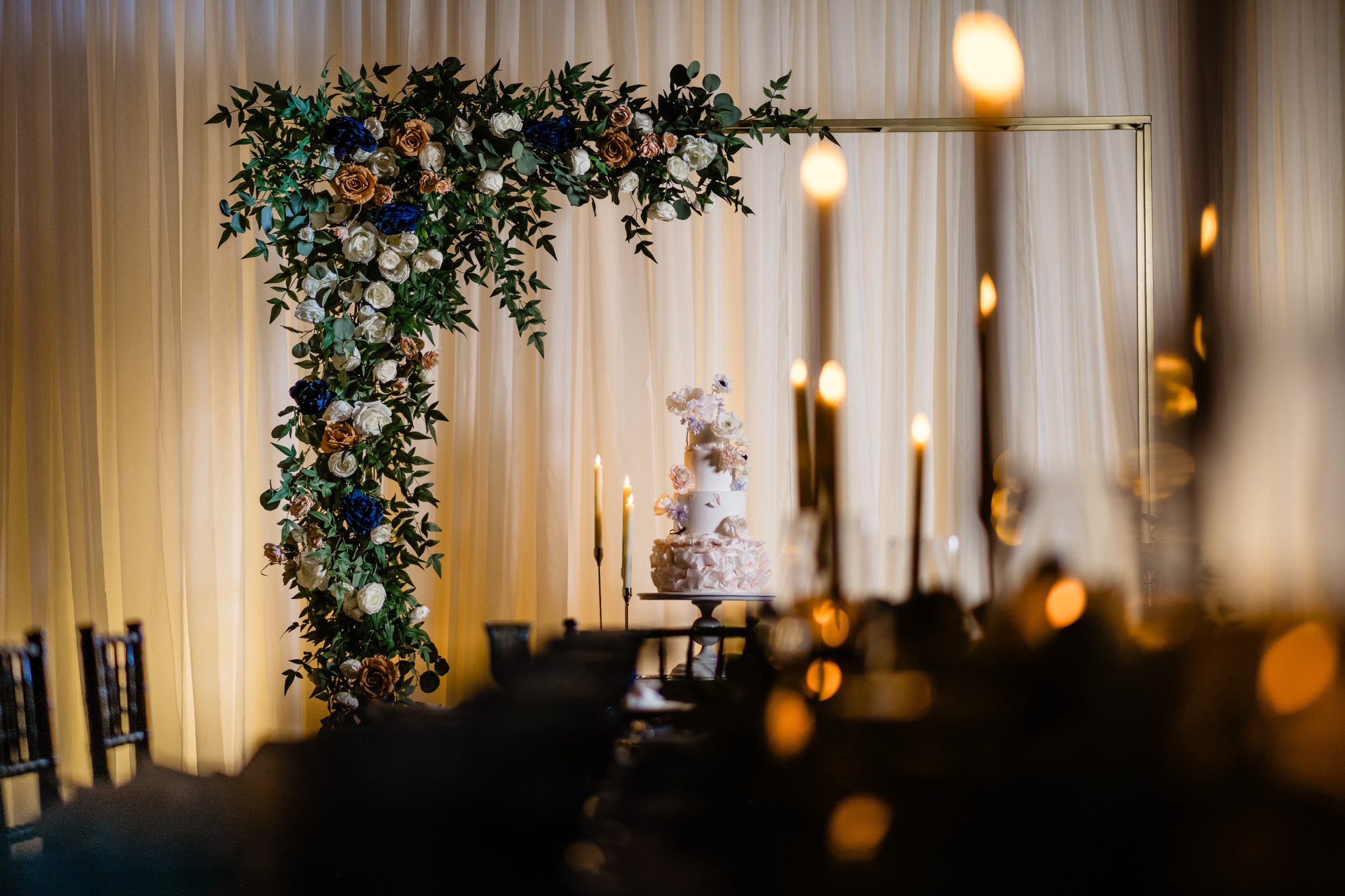 Decorative wedding arch adorned with flowers, with a wedding cake and candles in the background, against a beige curtain backdrop.