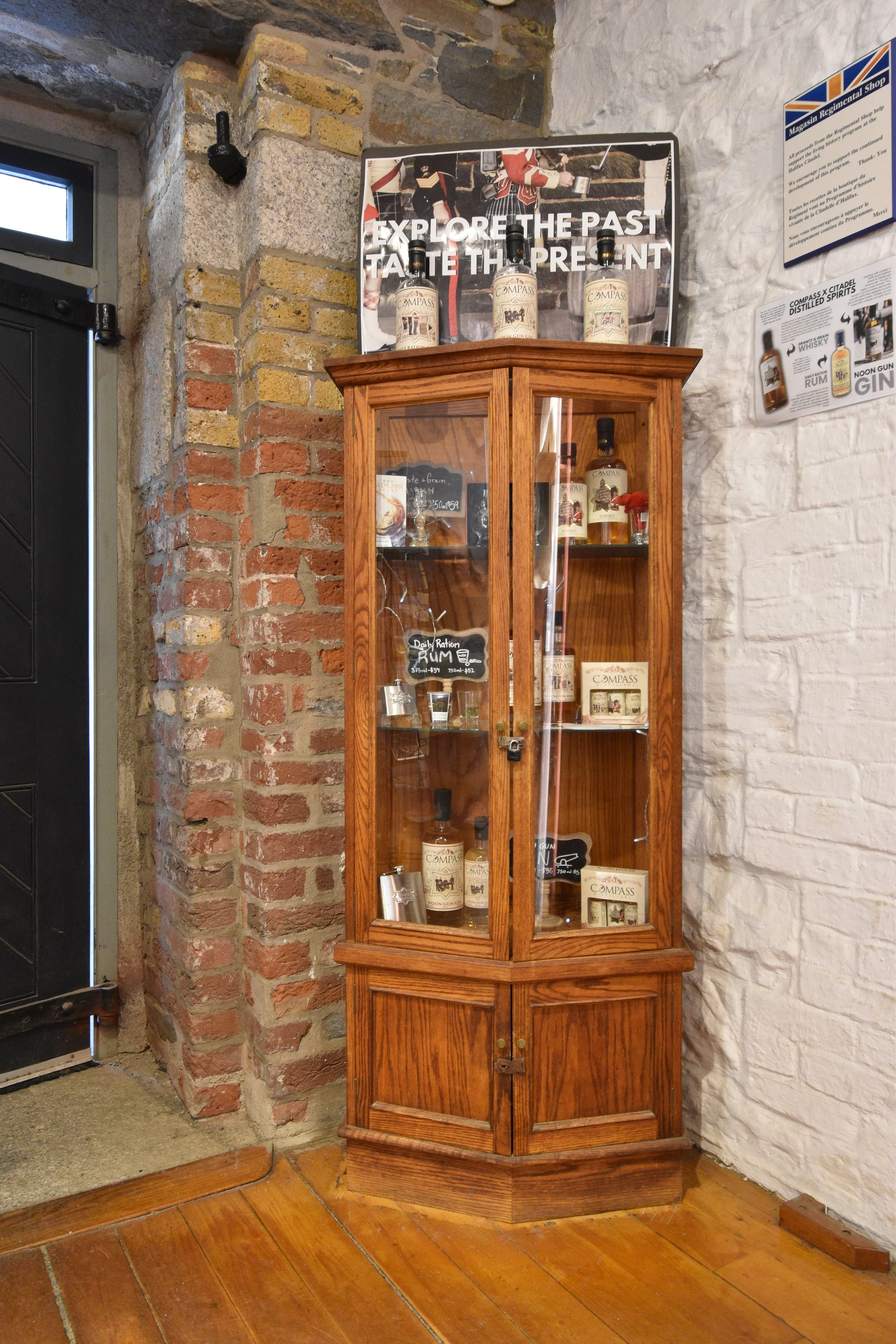 A wooden display cabinet in the Regitmental Shop. The cabinet showcases bottles of Compass brand spirits inside. The cabinet is positioned against a stone and brick wall, with a brick arch to the left and a white painted wall to the right.