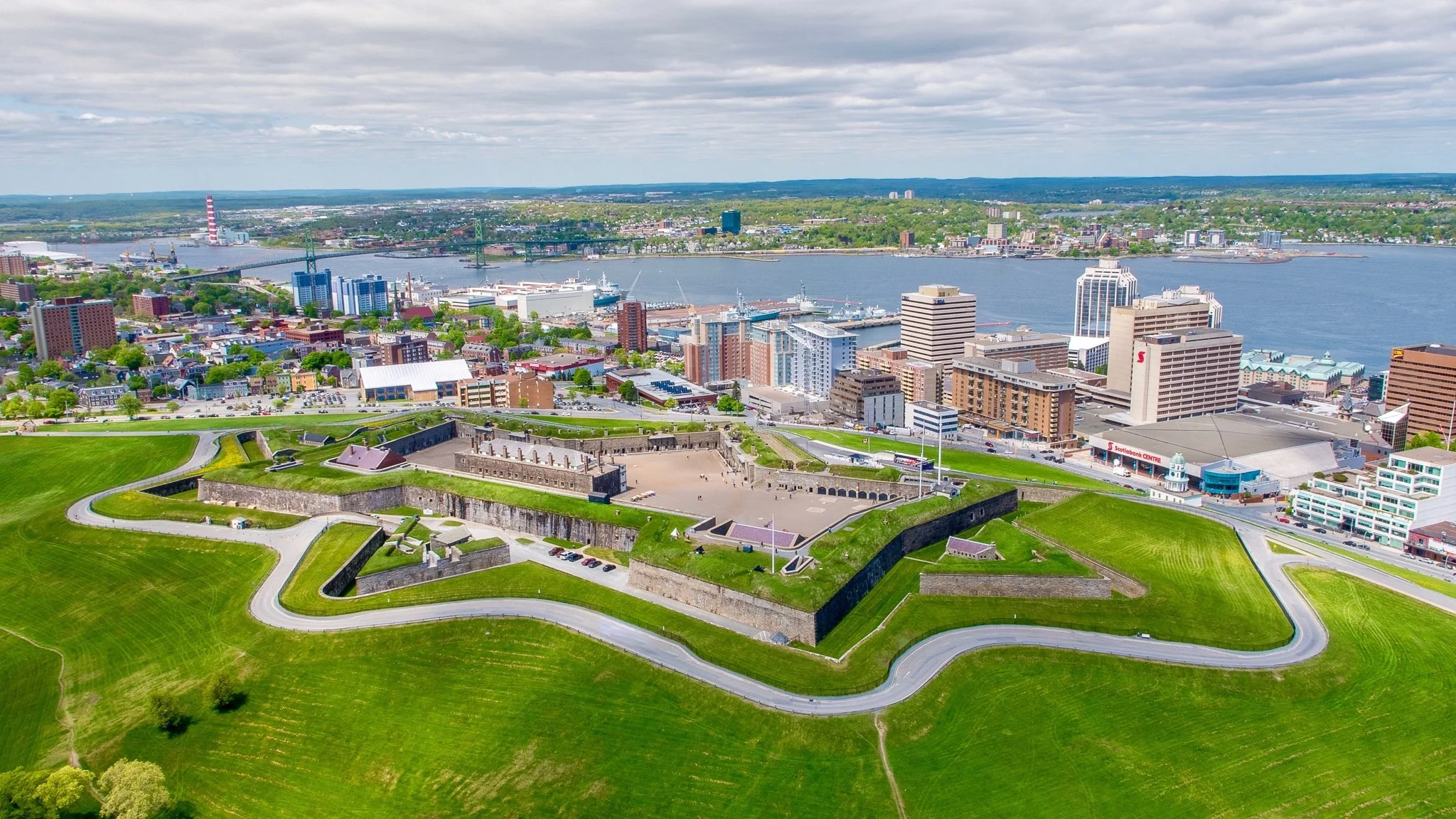 Aerial view of a city with a river running through it, featuring a mix of modern and historic buildings, a green park with fortress walls, and a bridge connecting different parts of the city under a cloudy sky.