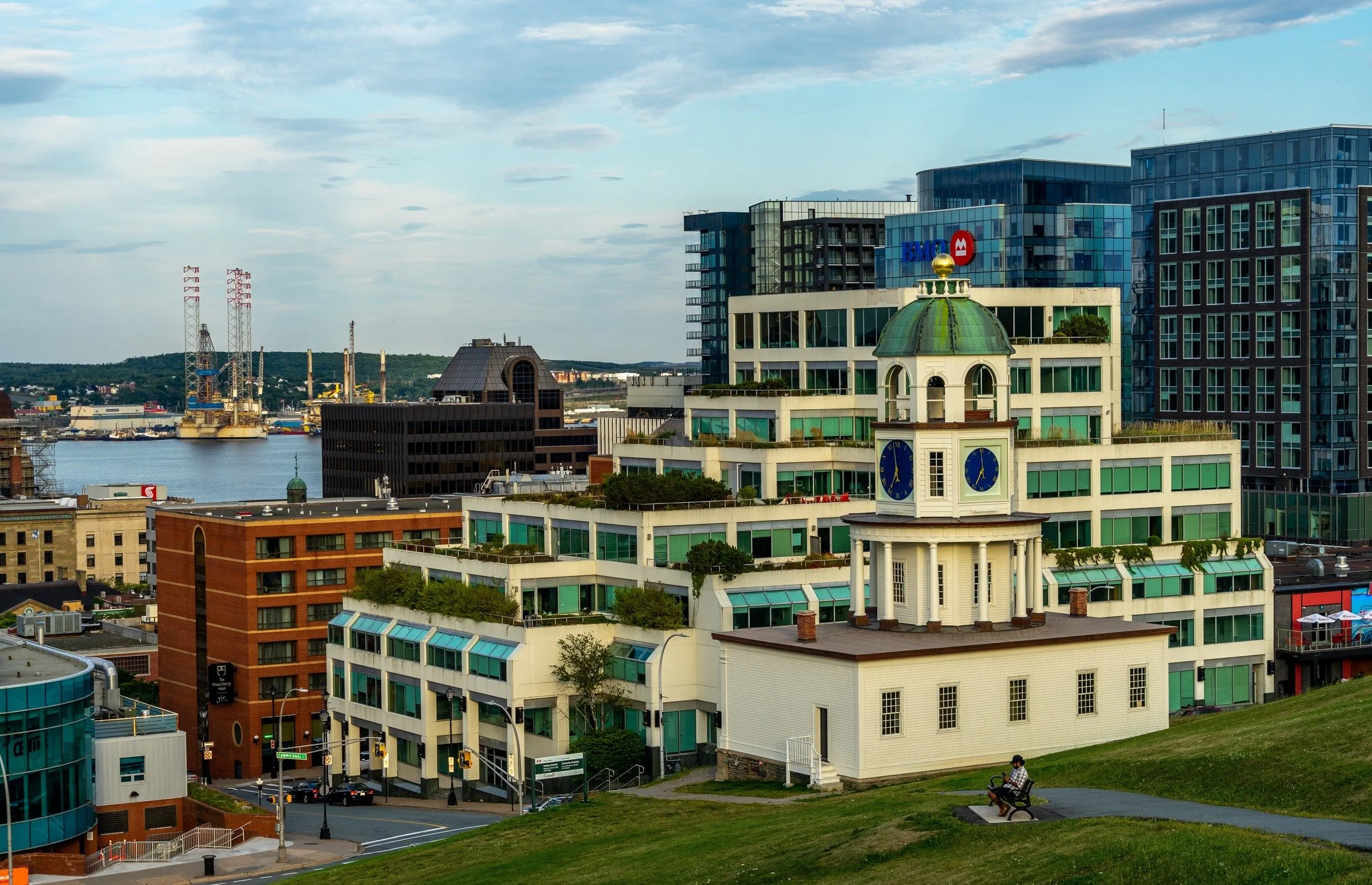 Cityscape with modern high-rise buildings, a historic clock tower, a river with industrial structures, and a person riding a bike in a park area.